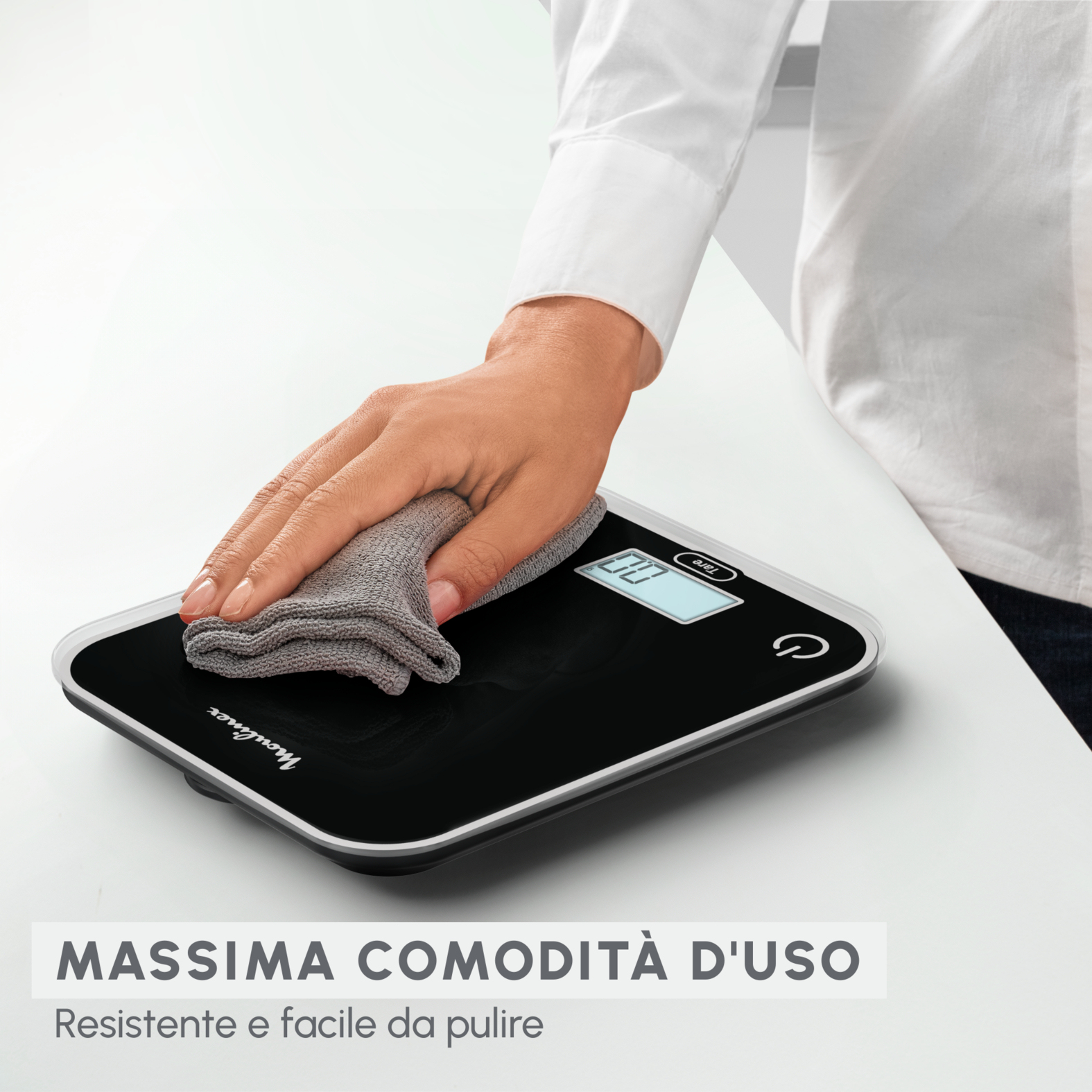 Dłoń wyciera czarną cyfrową wagę kuchenną szarą szmatką. Białe tło.