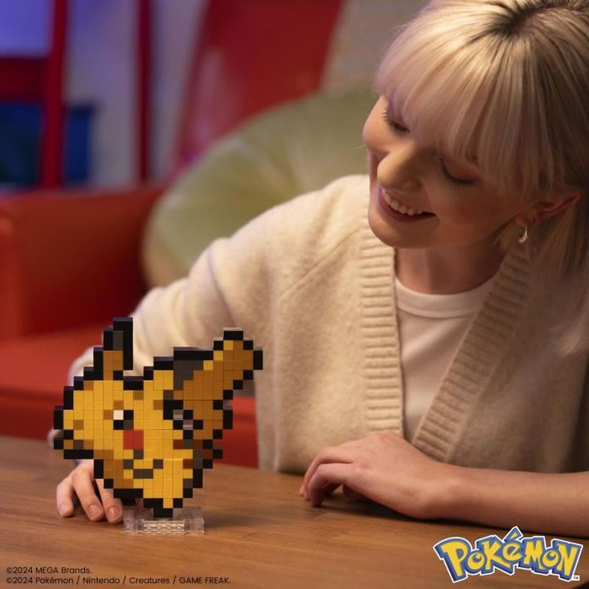 Une femme sourit en regardant une figurine Pikachu pixélisée sur une table en bois. Le logo Pokémon est visible.