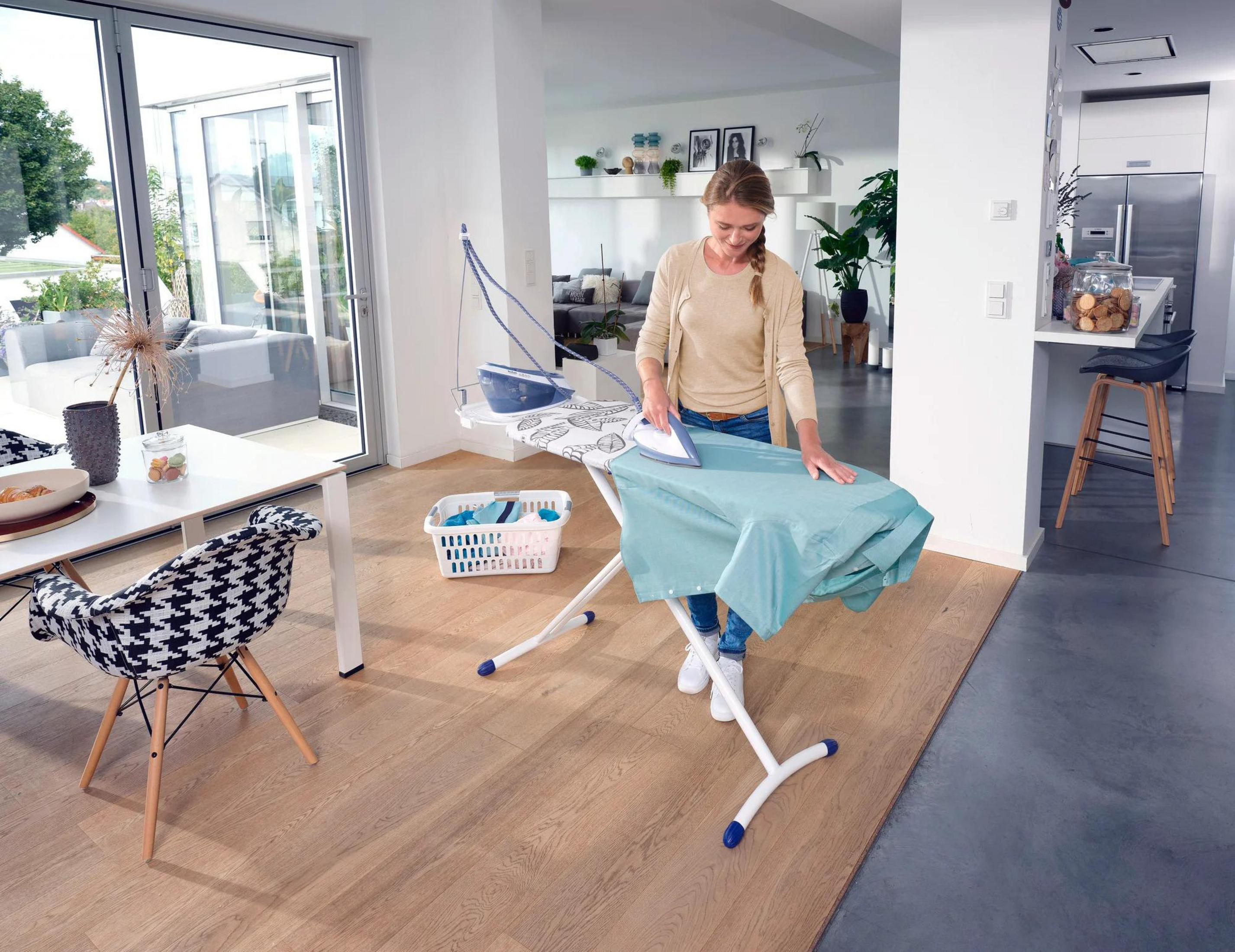 Kobieta prasuje turkusową koszulę na białej desce do prasowania. Pokój ma nowoczesny design z kuchnią i częścią dzienną.