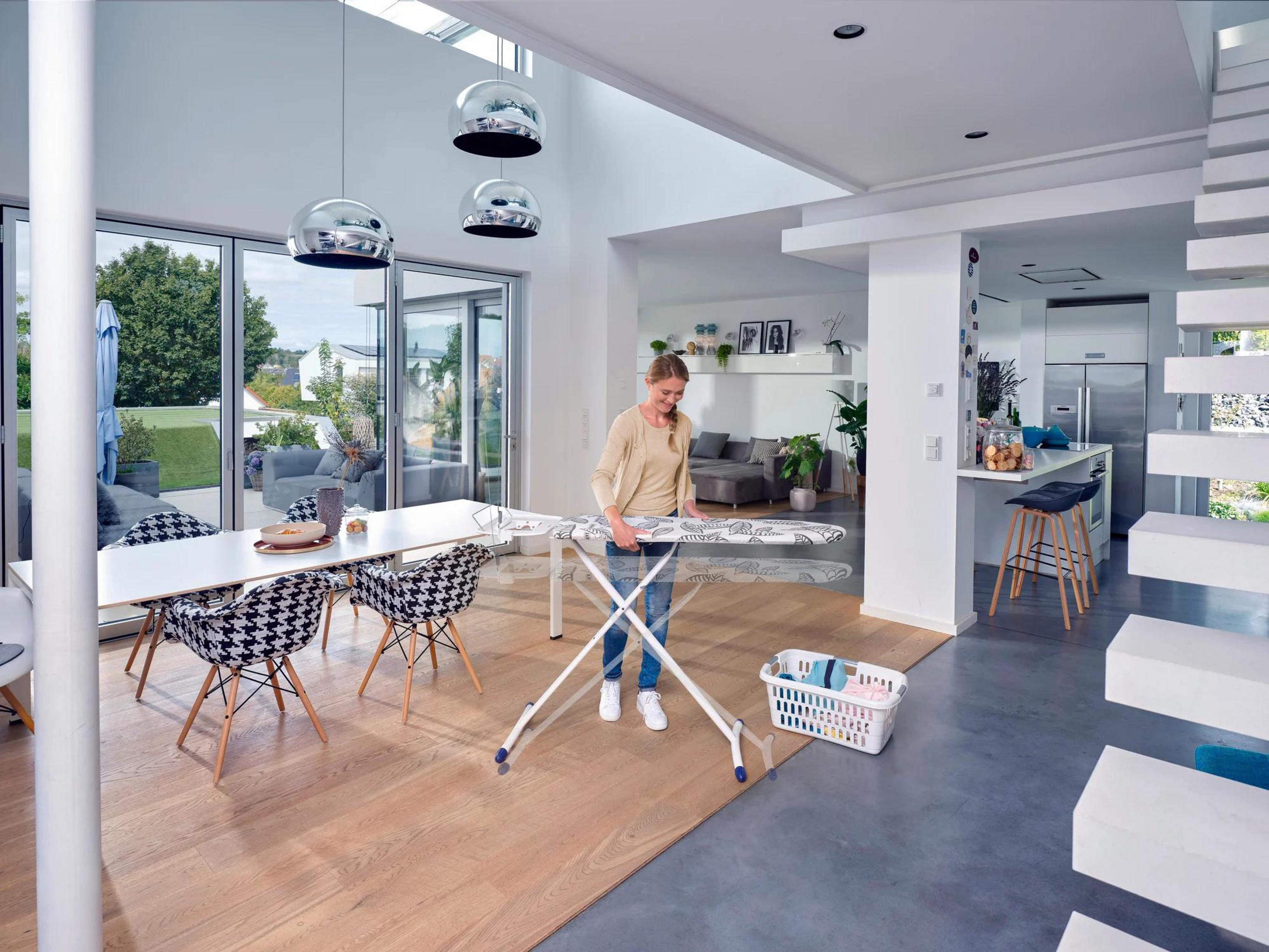 Kobieta prasuje na białej desce do prasowania. Pokój ma nowoczesny design z kuchnią i częścią dzienną. Białe meble.