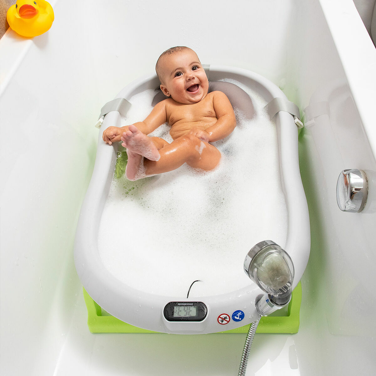 Baby in Badewanne mit Blasen, lächelnd. Eine gelbe Quietscheente liegt daneben.