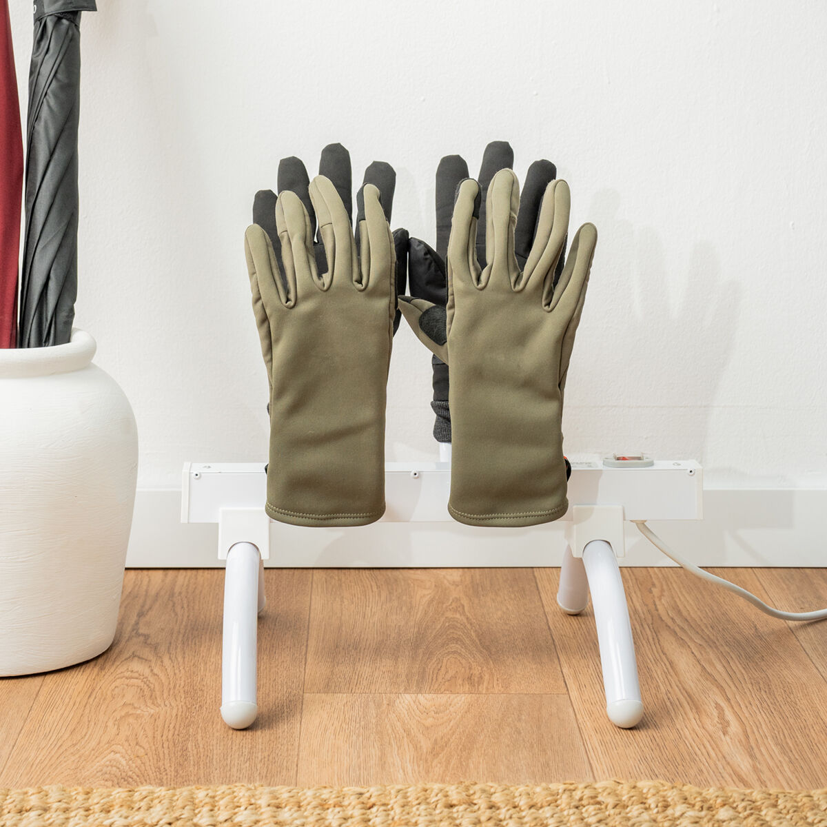 Un estante blanco con guantes encima se encuentra sobre un suelo de madera junto a un jarrón blanco y paraguas.