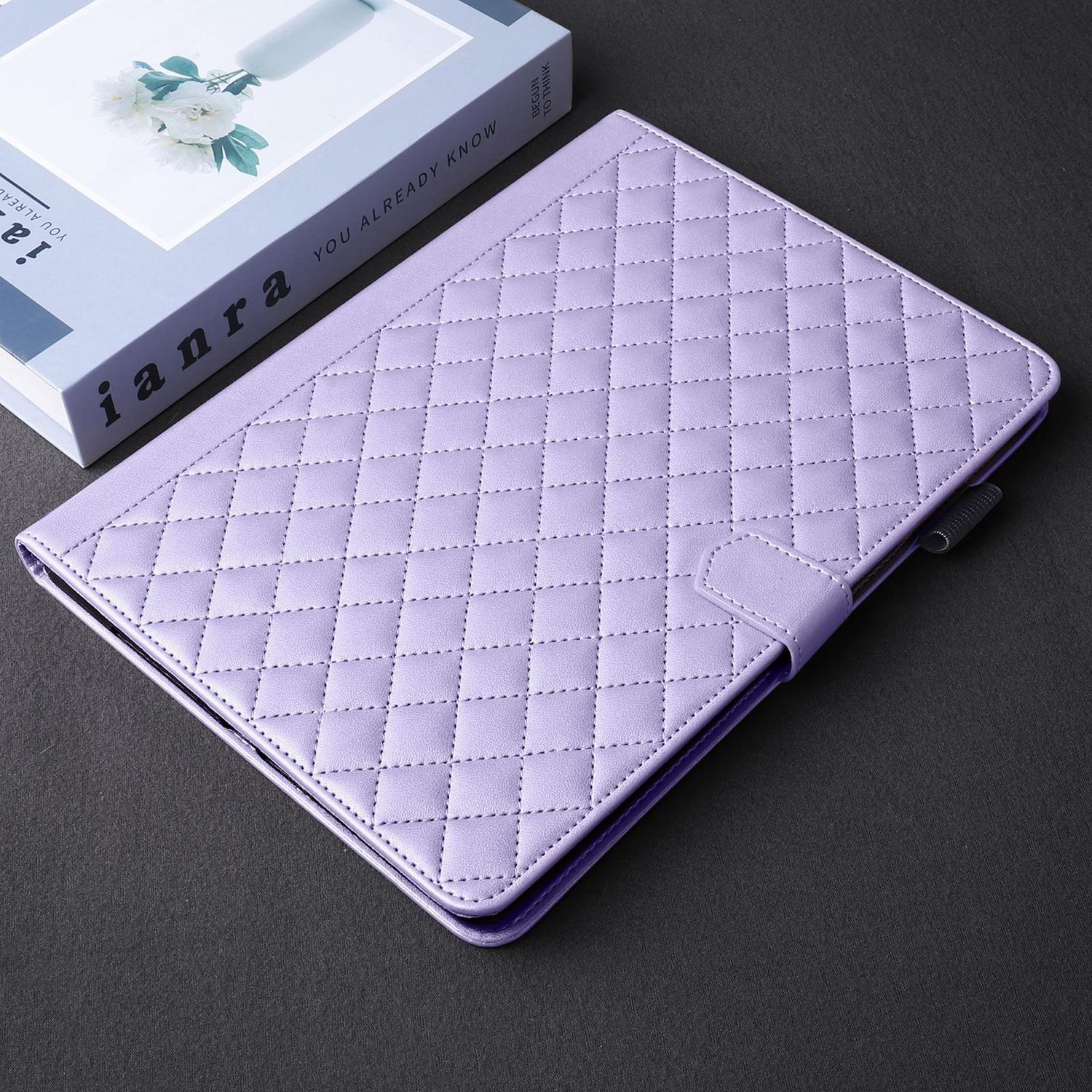 Lila Tablet-Hülle auf einem Tisch mit einem Buch. Die Hülle ist geschlossen. Der Hintergrund ist dunkel.
