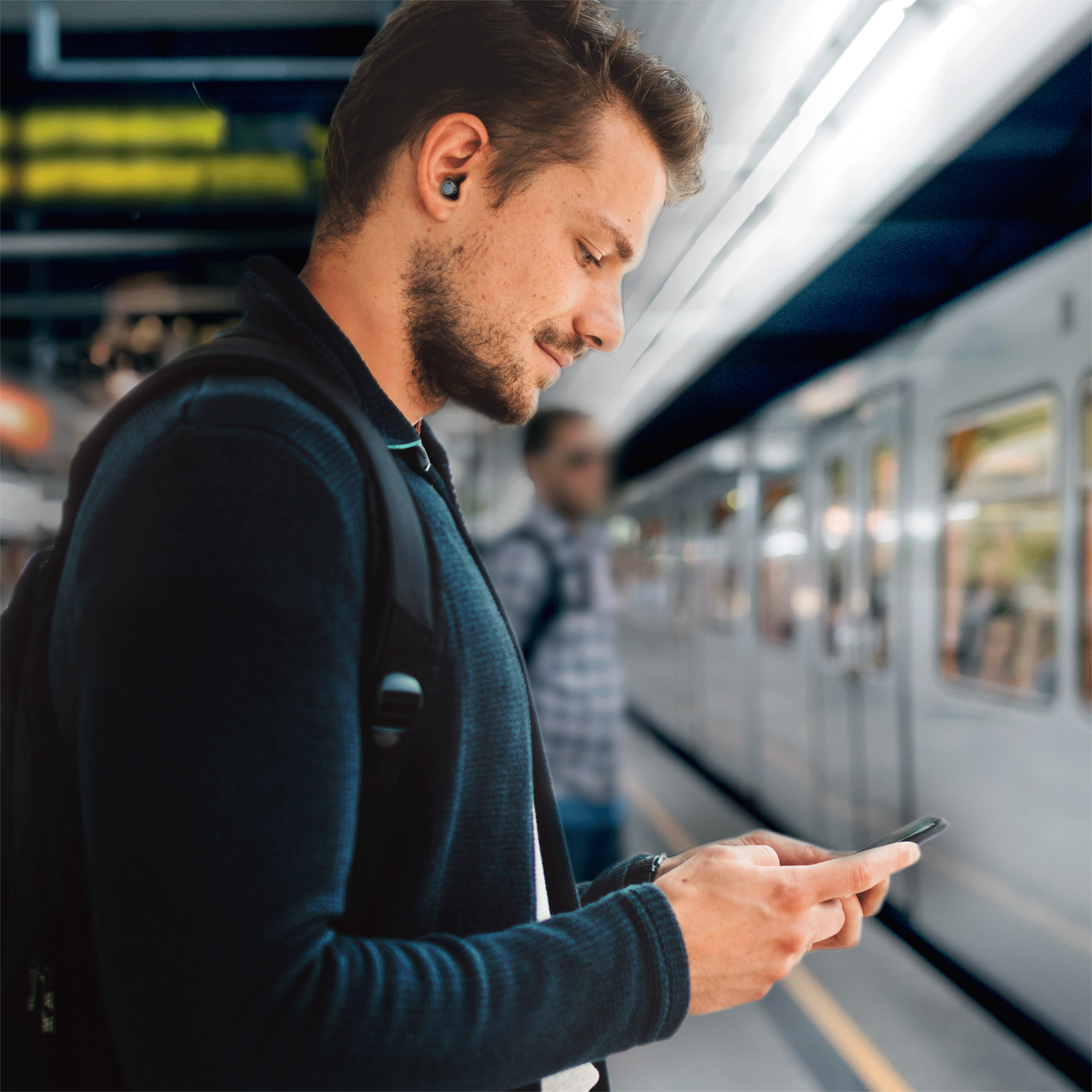 Een man met oordopjes die een telefoon gebruikt terwijl hij op een treinstation staat. Een trein is zichtbaar op de achtergrond.