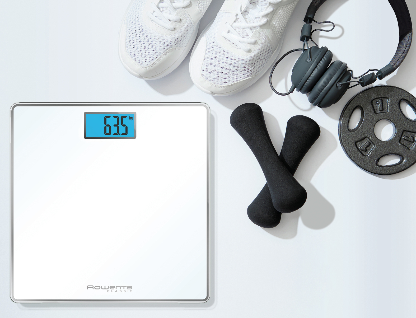 Una bilancia digitale, scarpe da ginnastica, manubri e cuffie sono disposti su una superficie bianca. La bilancia mostra 63,5 kg.