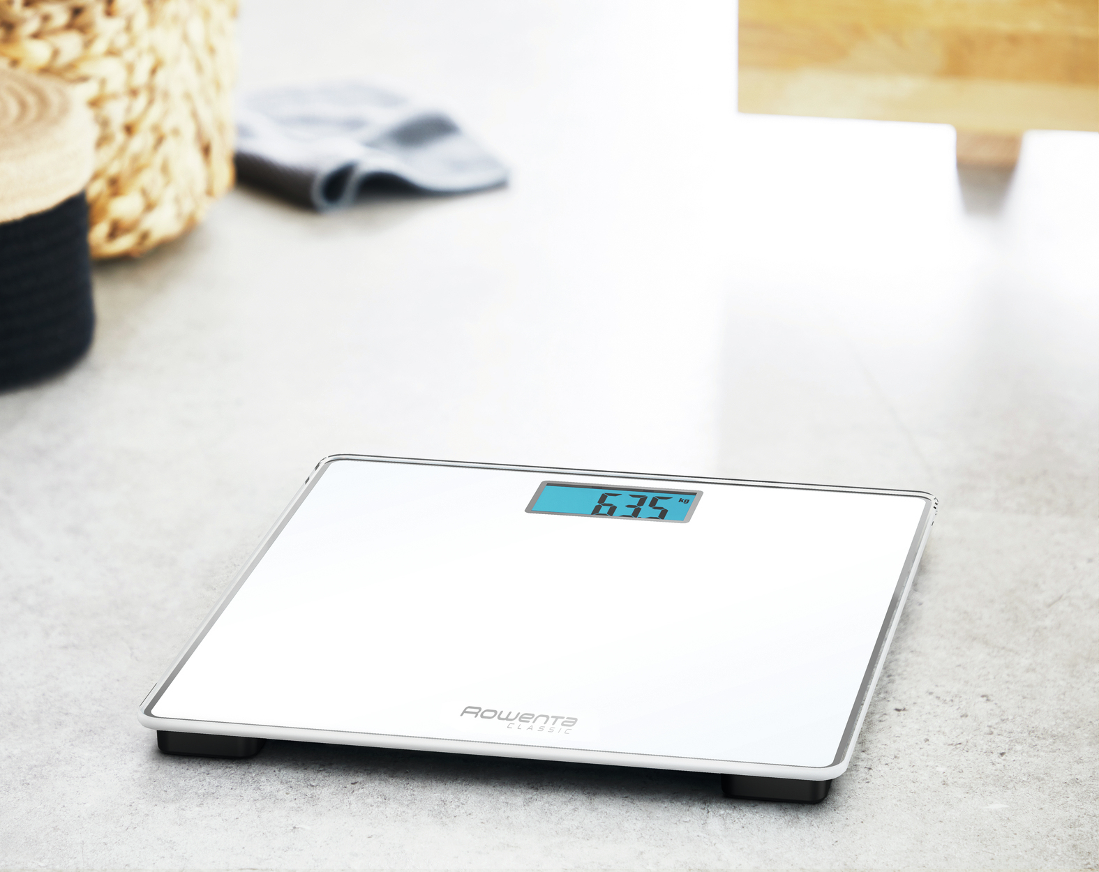 Bilancia da bagno digitale bianca. Il display mostra 63,5 kg. Cestino in vimini e asciugamano sullo sfondo.