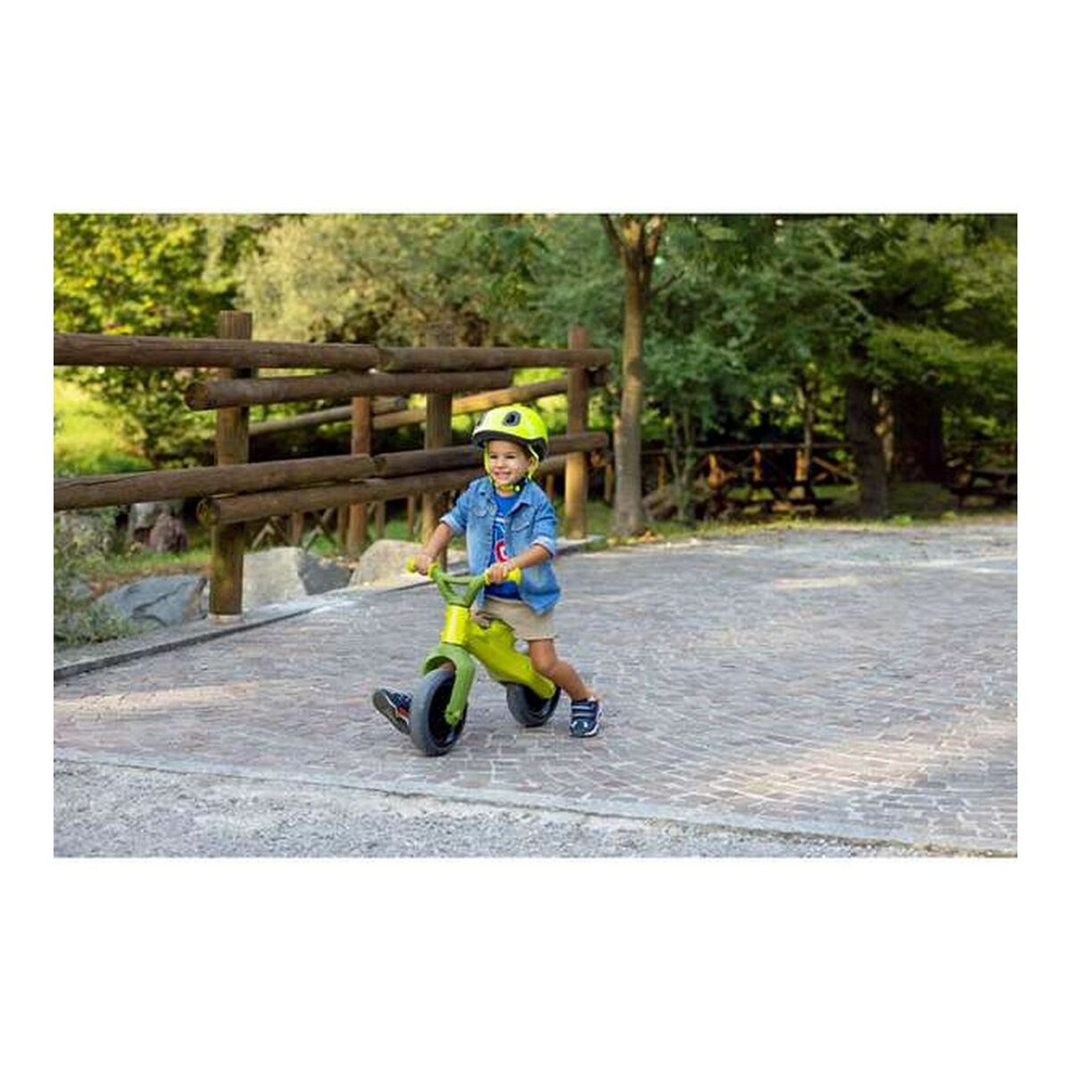 Un bambino con un casco che va in bici senza pedali verde su un sentiero pavimentato, vicino a una staccionata di legno.
