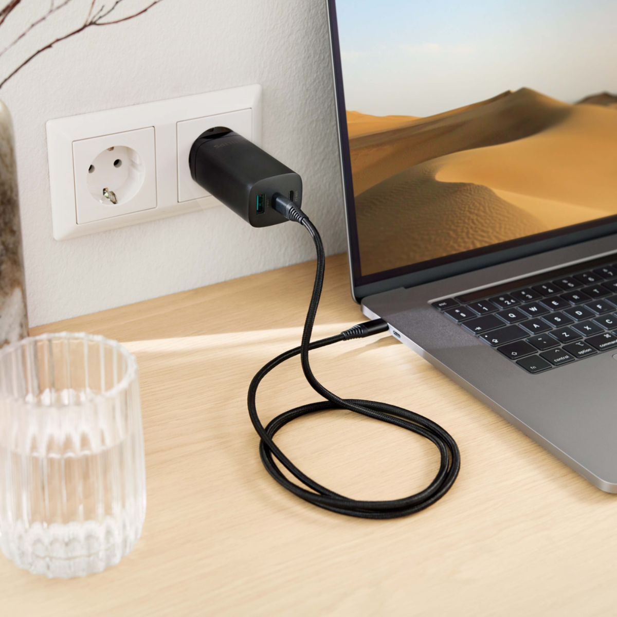 Laptop charging via USB-C charger plugged into a wall outlet. A glass of water is on the table.