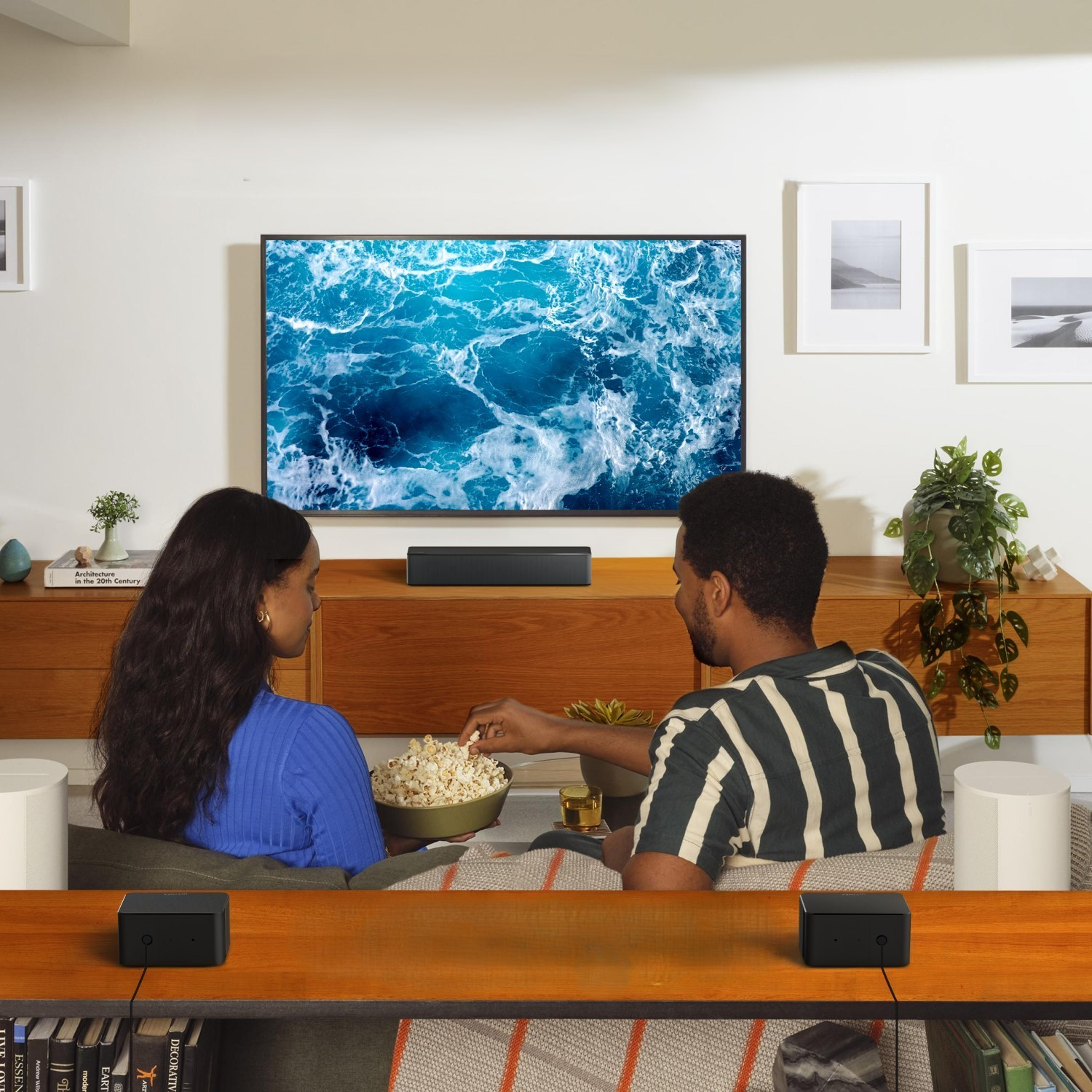 Dwie osoby oglądają telewizję i jedzą popcorn. W pokoju znajduje się sofa, głośniki i drewniana szafka.