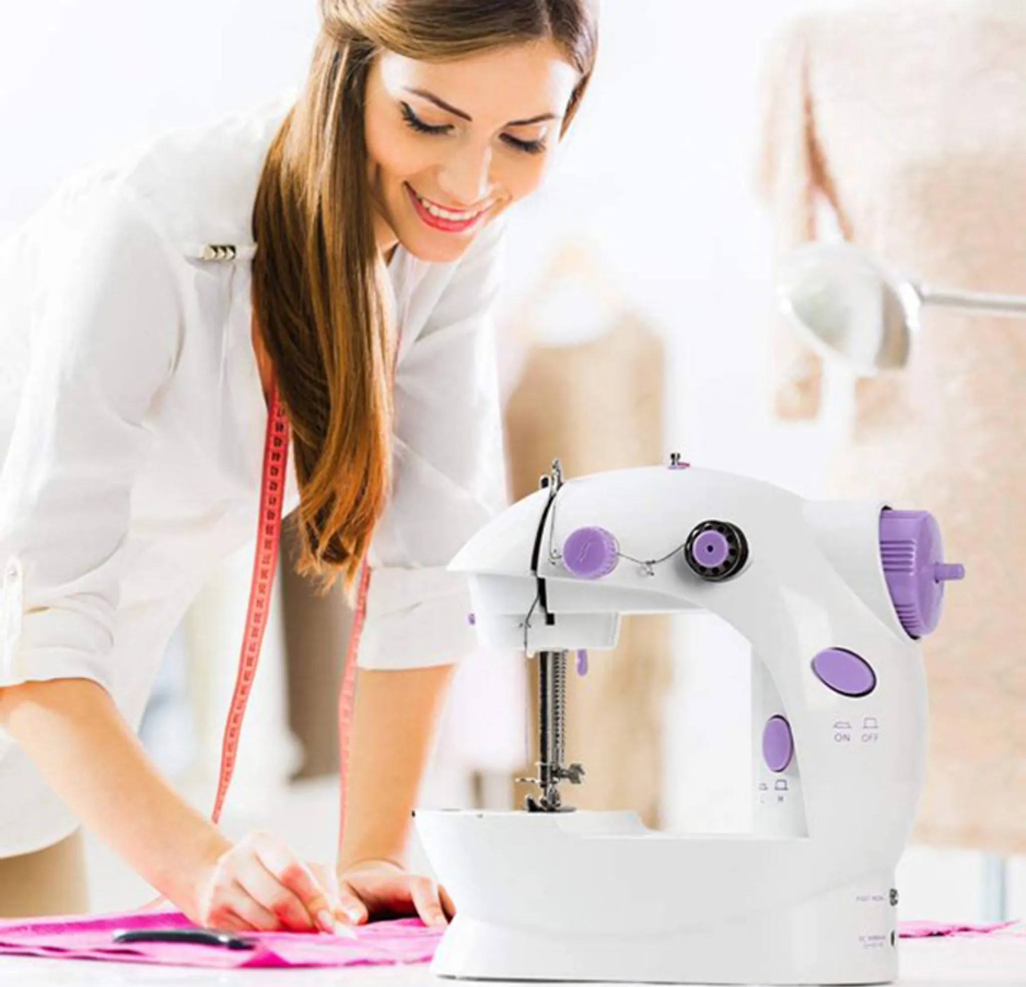 Frau lächelt, näht mit weißer Maschine. Rosa Stoff auf dem Tisch. Hintergrund: Schneiderpuppe im verschwommenen Raum.