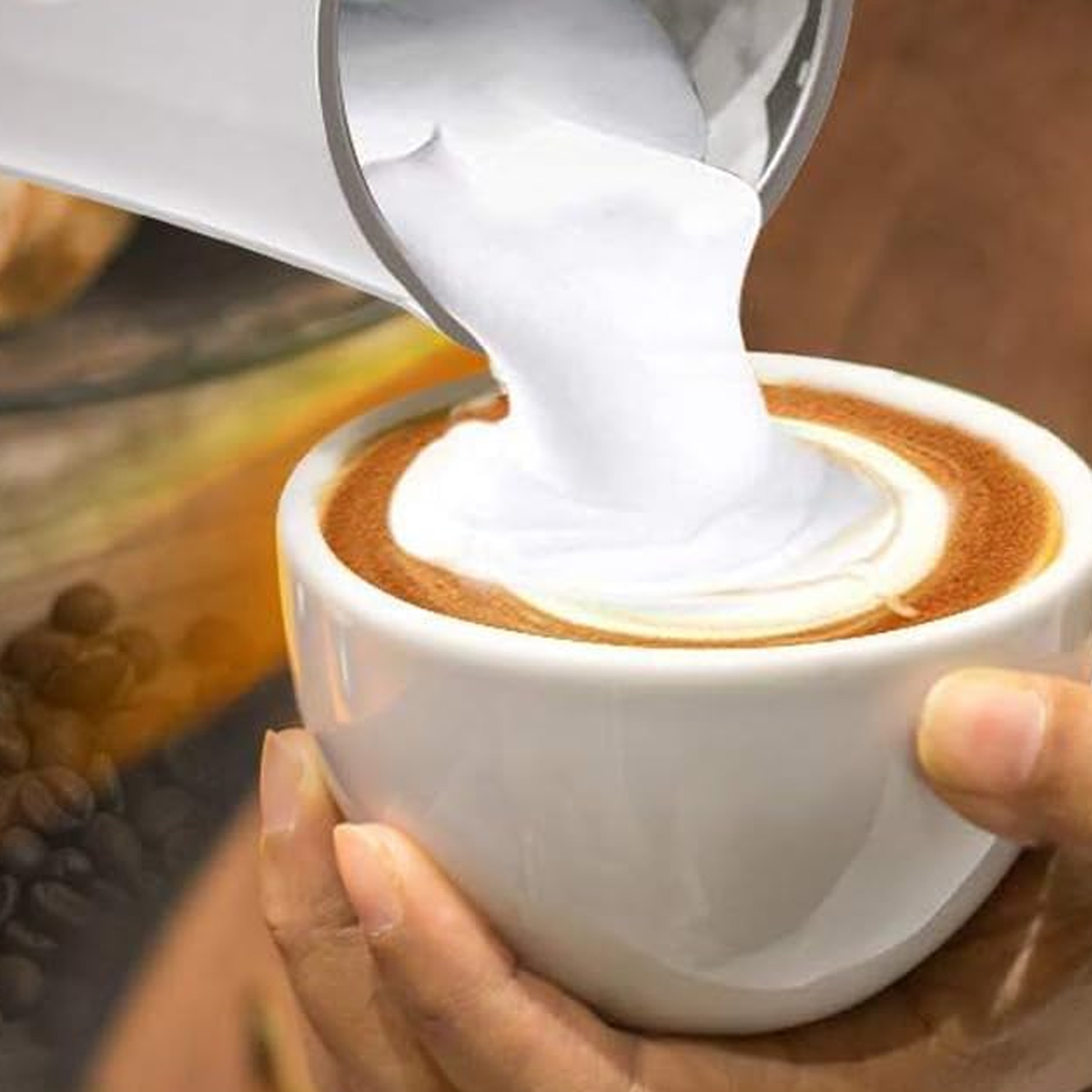 Milch wird in eine Kaffeetasse mit Latte Art gegossen. Hand hält die Tasse.