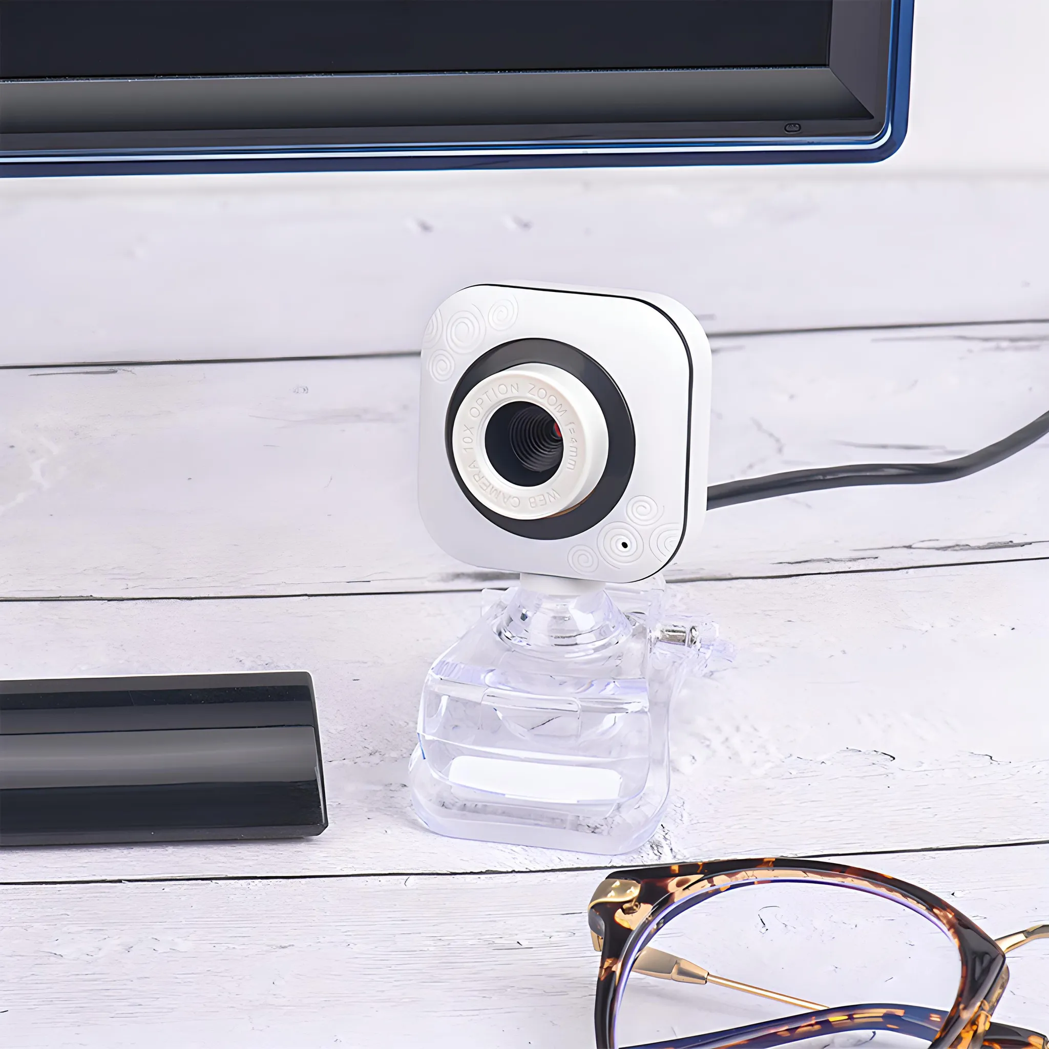 Eine weiße Webcam auf einem Schreibtisch mit Brille vor einem Computerbildschirm.