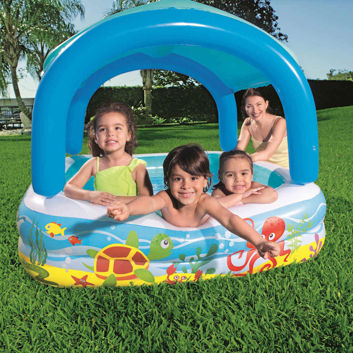 Enfants dans une piscine avec un auvent. Une femme est en arrière-plan. Créatures marines colorées sont sur le côté de la piscine.