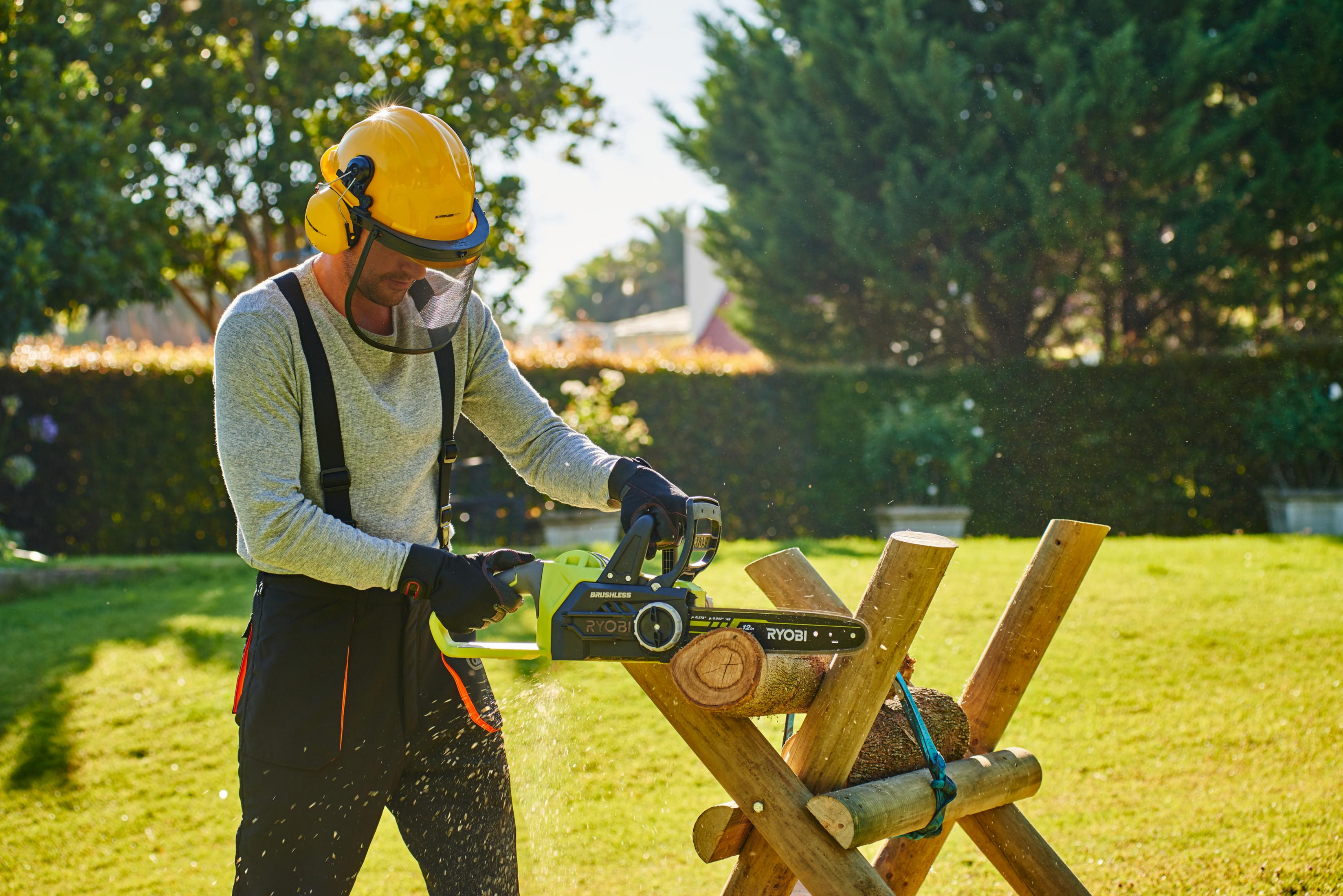 Mężczyzna w sprzęcie ochronnym używa piły łańcuchowej do cięcia kłody na koziołku na zewnątrz.