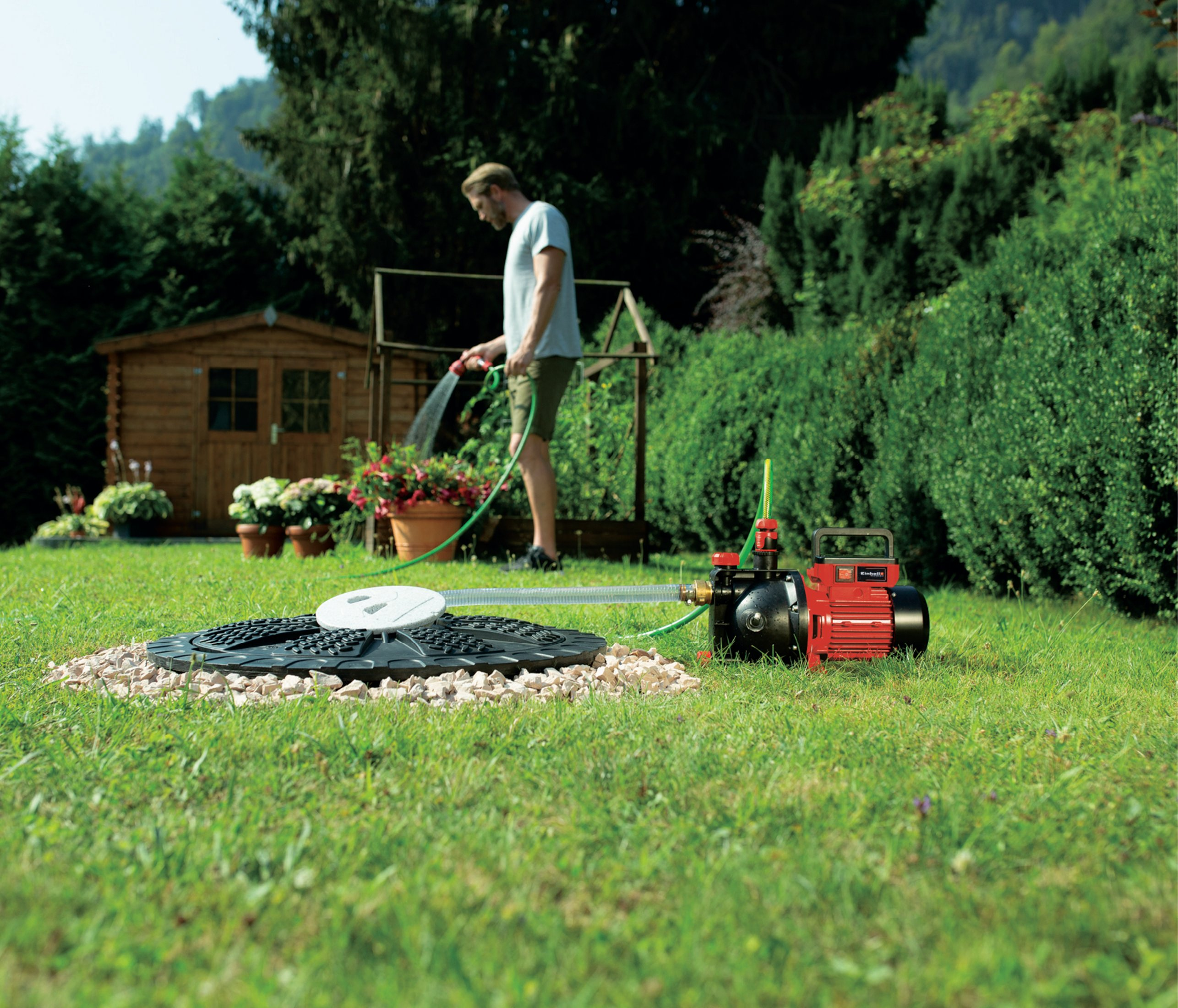 Mężczyzna podlewający rośliny wężem w ogrodzie z pompą. Zielony trawnik i drzewa.