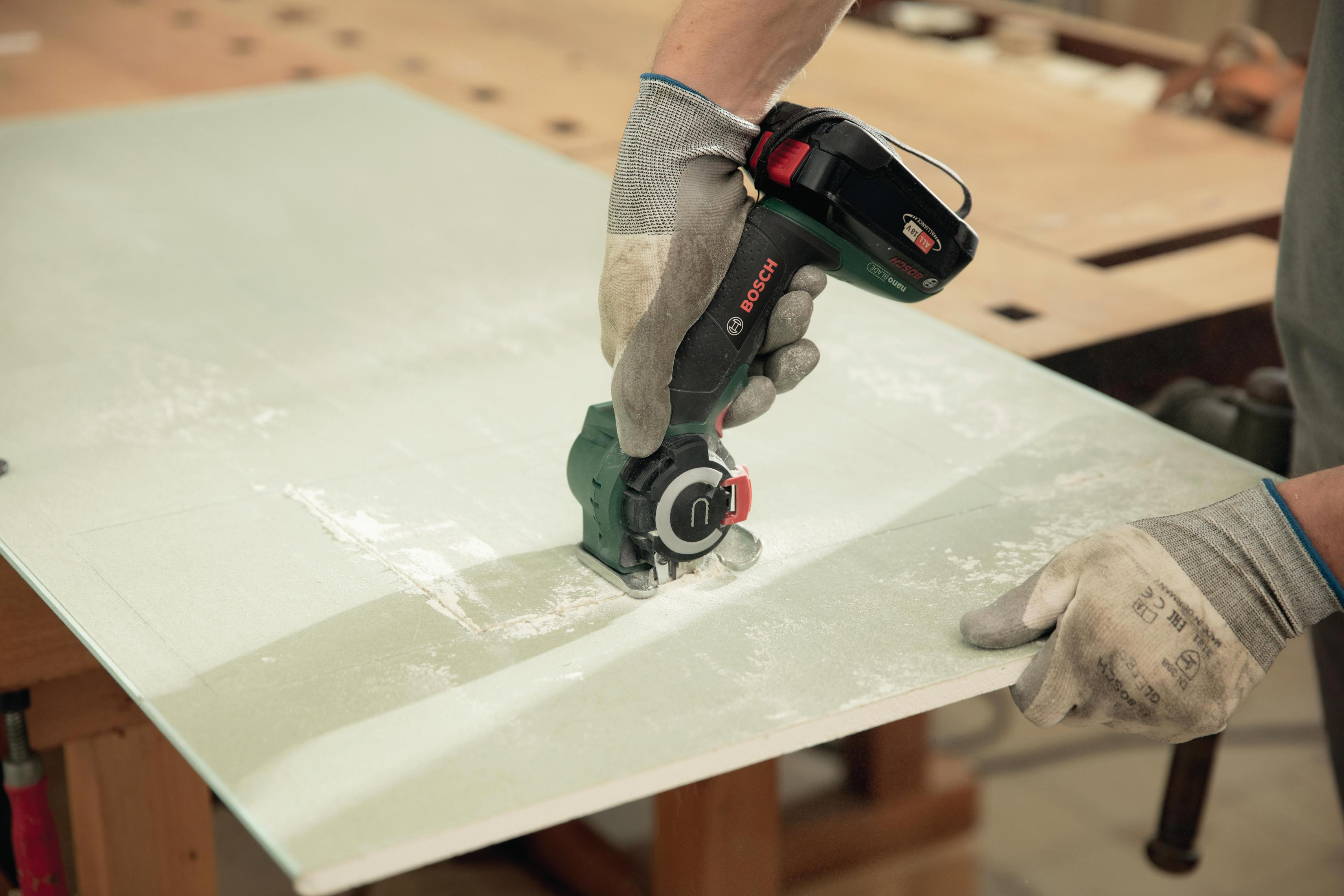 Ręce trzymają mini piłę tarczową Bosch, tnąc płytę gipsowo-kartonową.
