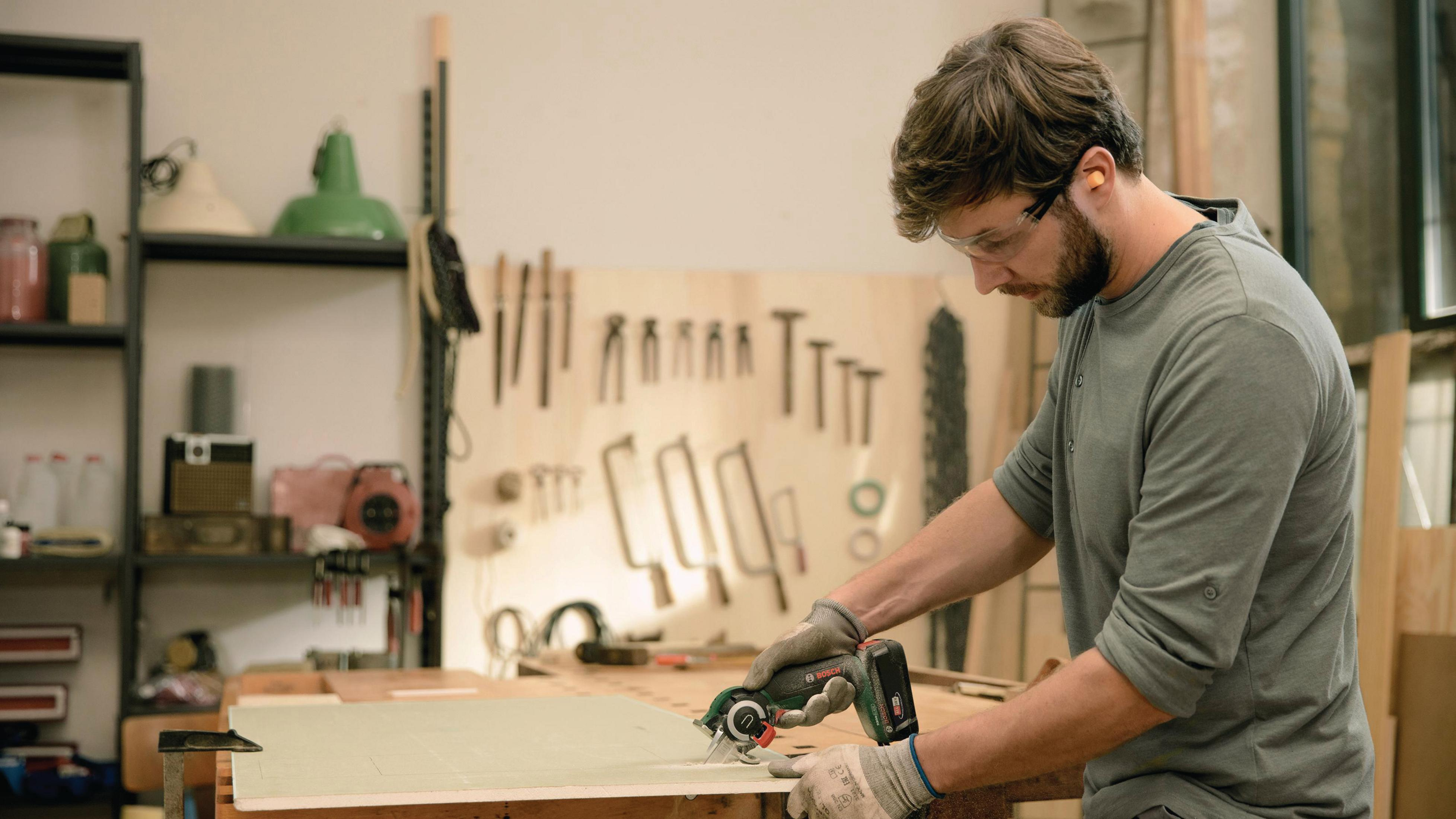 Mężczyzna używa piły elektrycznej do cięcia kawałka drewna. Nosi okulary ochronne i ochronniki słuchu.