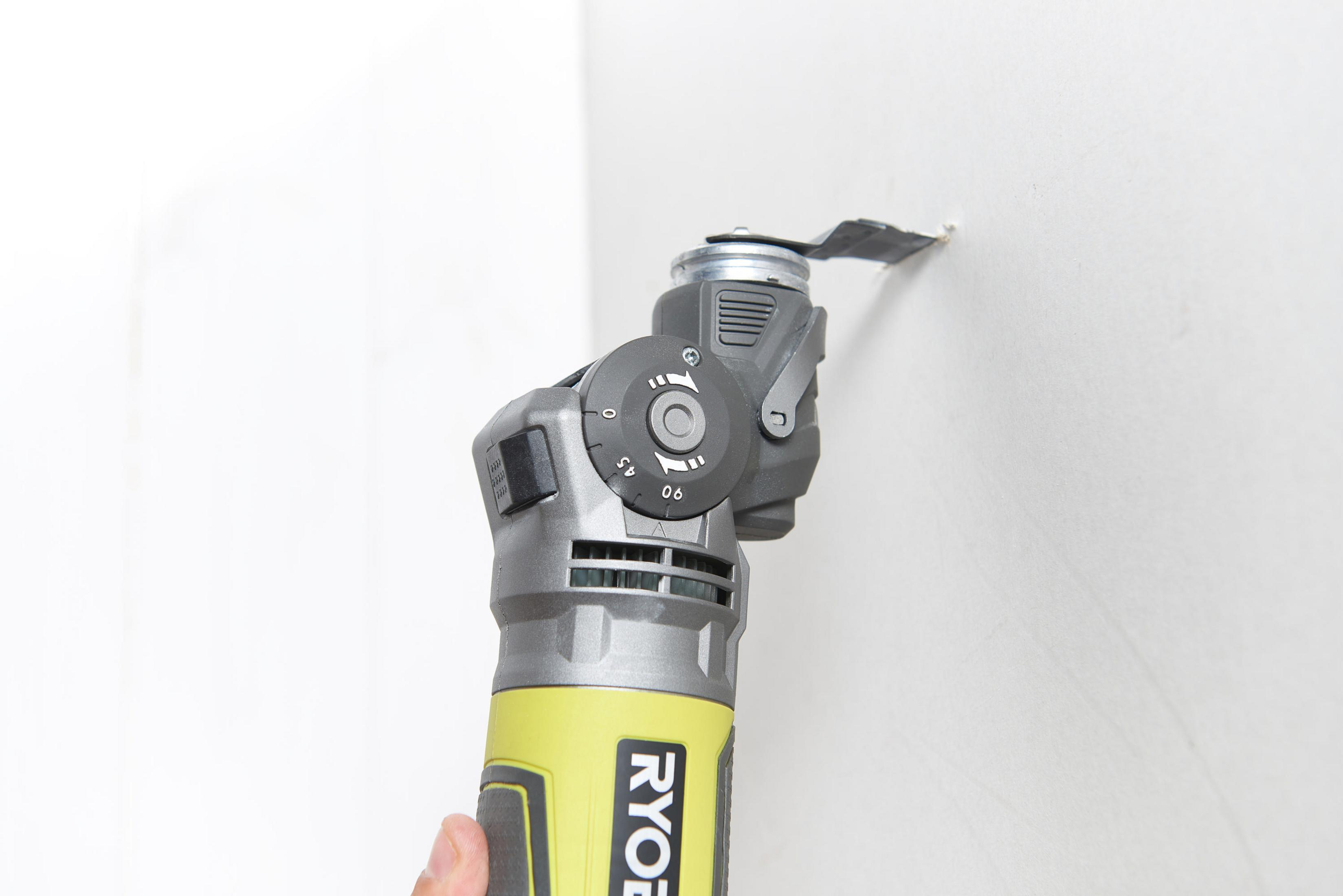 Grünes und graues Oszillationswerkzeug schneidet in eine weiße Wand. RYOBI ist auf der Seite des Werkzeugs aufgedruckt.