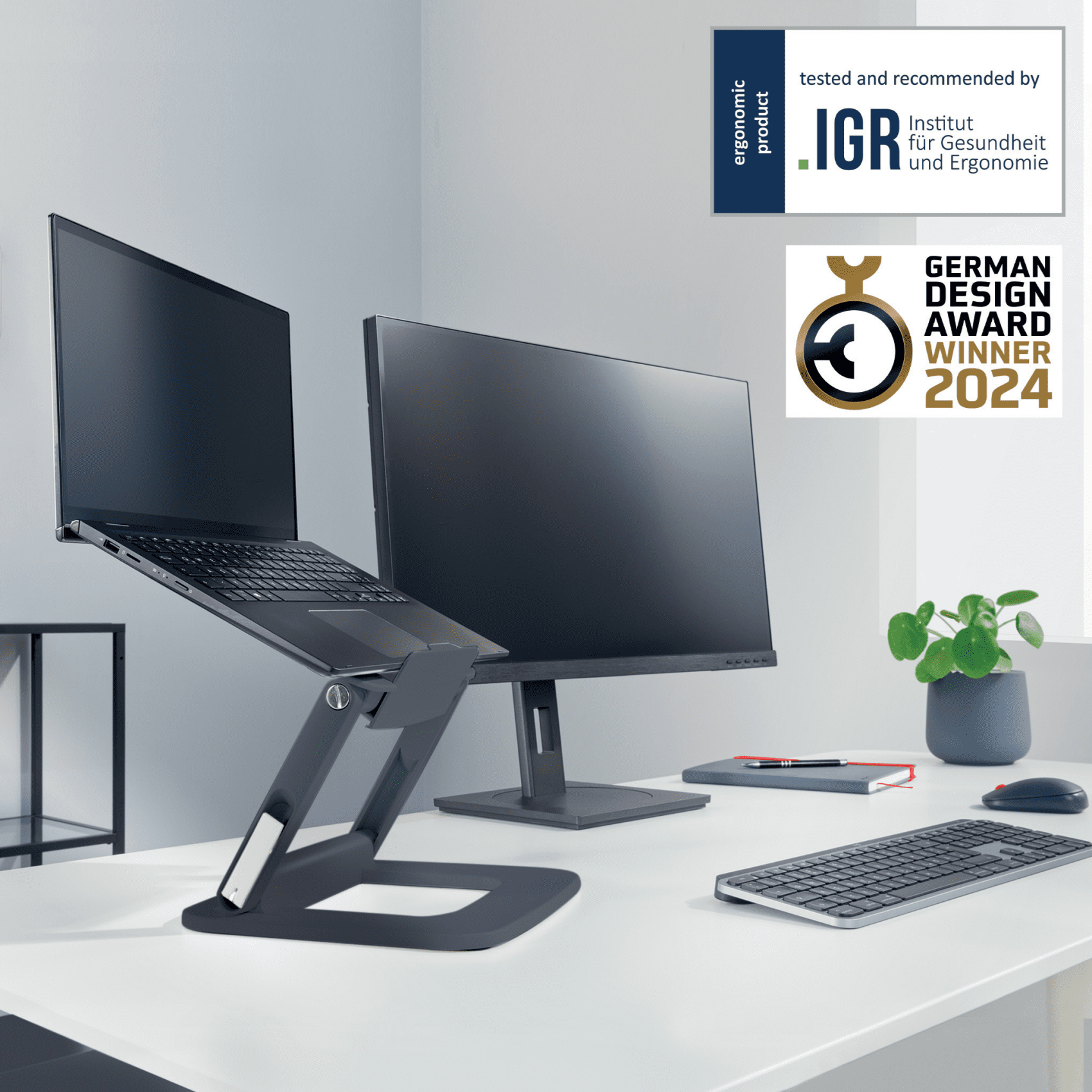 Ein Laptop und ein Monitor auf einem Ständer auf einem weißen Schreibtisch. Auszeichnungen und eine Pflanze sind sichtbar.
