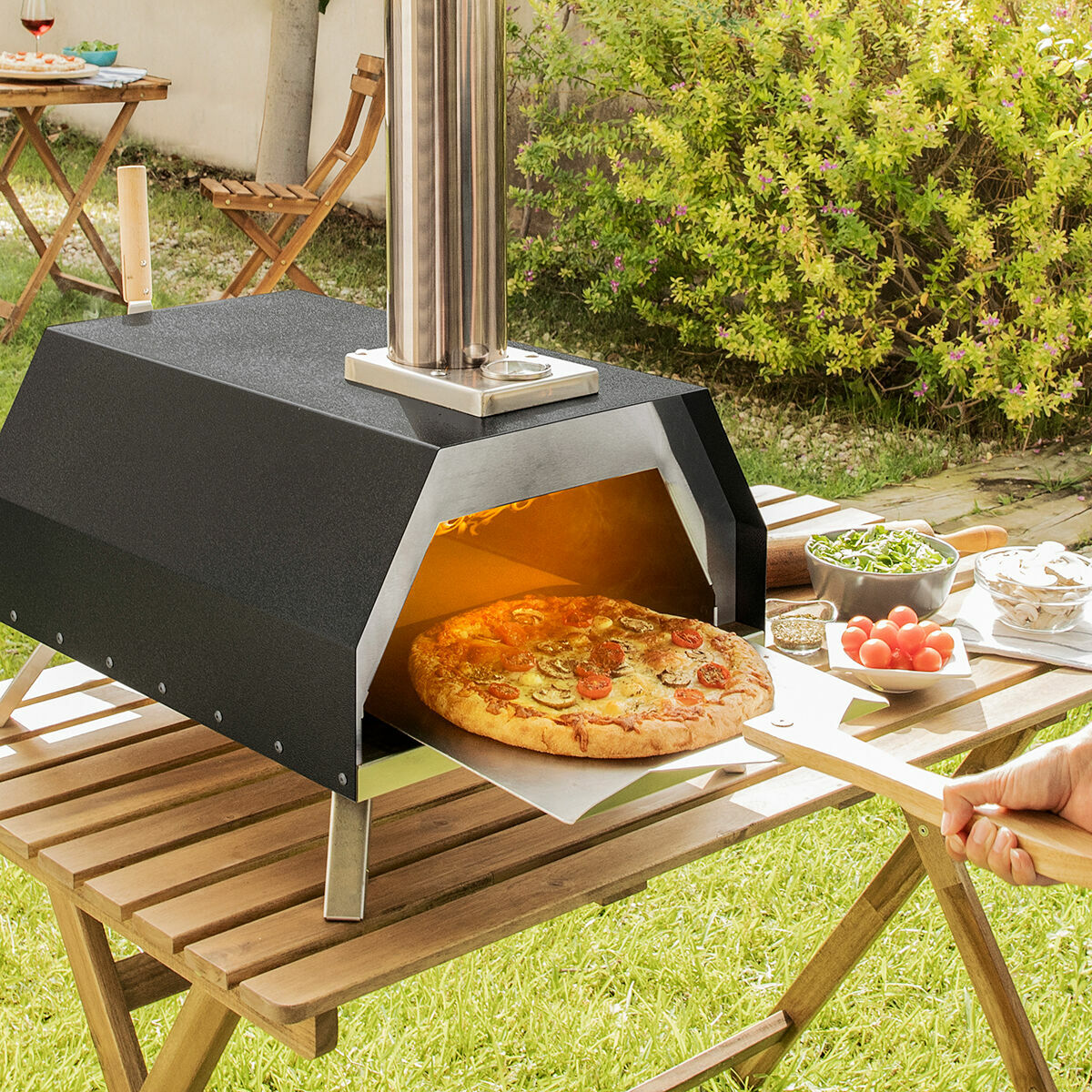 Pizza in un forno per pizza nero all'aperto, su un tavolo di legno e ingredienti.