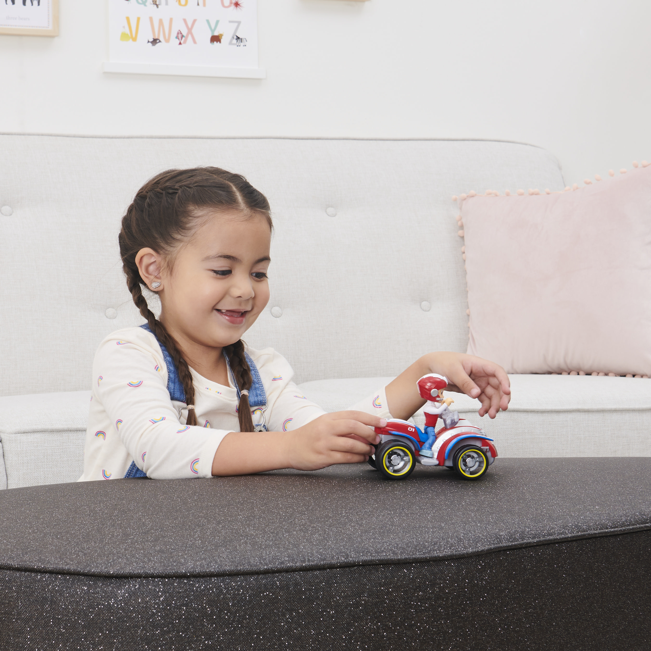 Una niña está jugando con una figura de juguete Ryder y un ATV. La niña está sonriendo. Un sofá está en el fondo.