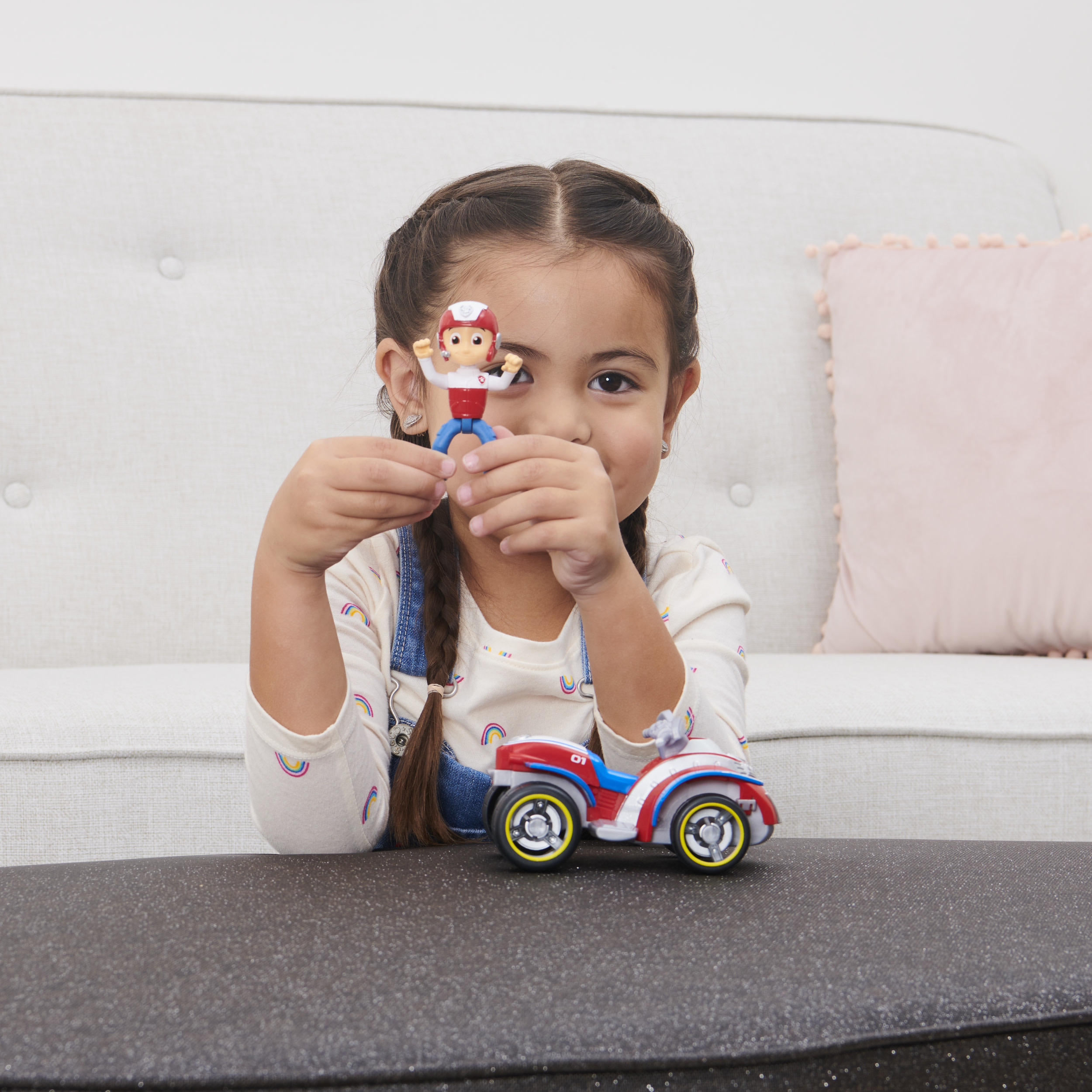 Una niña sostiene una figura de juguete Ryder y un ATV. Un sofá y una superficie gris oscuro están al fondo.