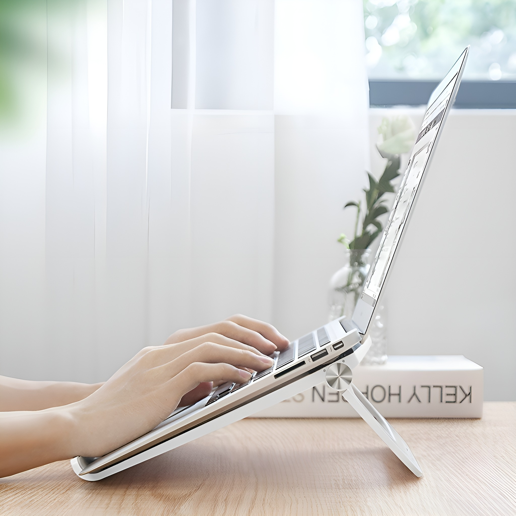 Eine Person benutzt einen Laptop auf einem weißen Ständer auf einem Holztisch in der Nähe eines Fensters.