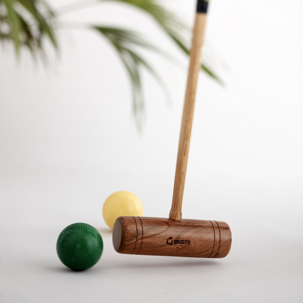 Ein Krocket-Schläger und zwei Bälle, einer gelb und einer grün, auf einer weißen Oberfläche mit Palmenblatt.