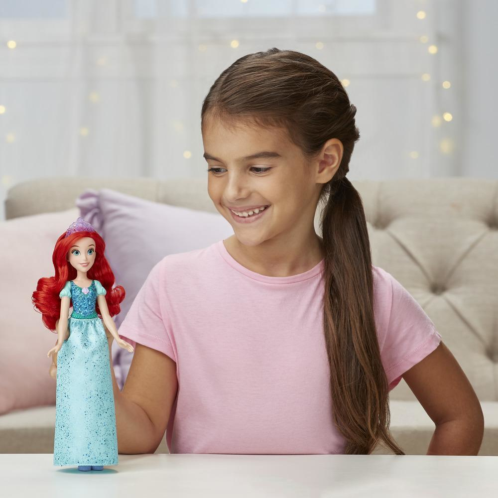Ragazza tiene bambola Ariel. Ariel ha capelli rossi, vestito blu. Sfondo: divano beige.