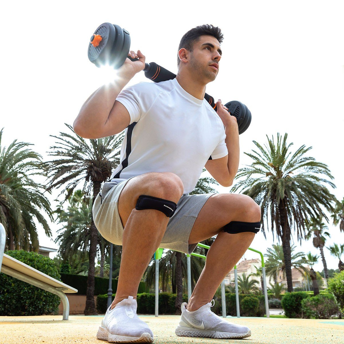 Uomo in posizione squat, con pesi. Ginocchiere, scarpe bianche. Palme sullo sfondo.