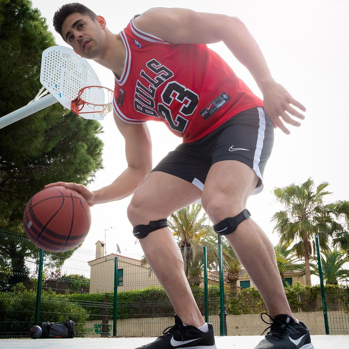 Uomo che gioca a basket, con maglia e pantaloncini. Ginocchiere, scarpe da ginnastica. Campo con rete.