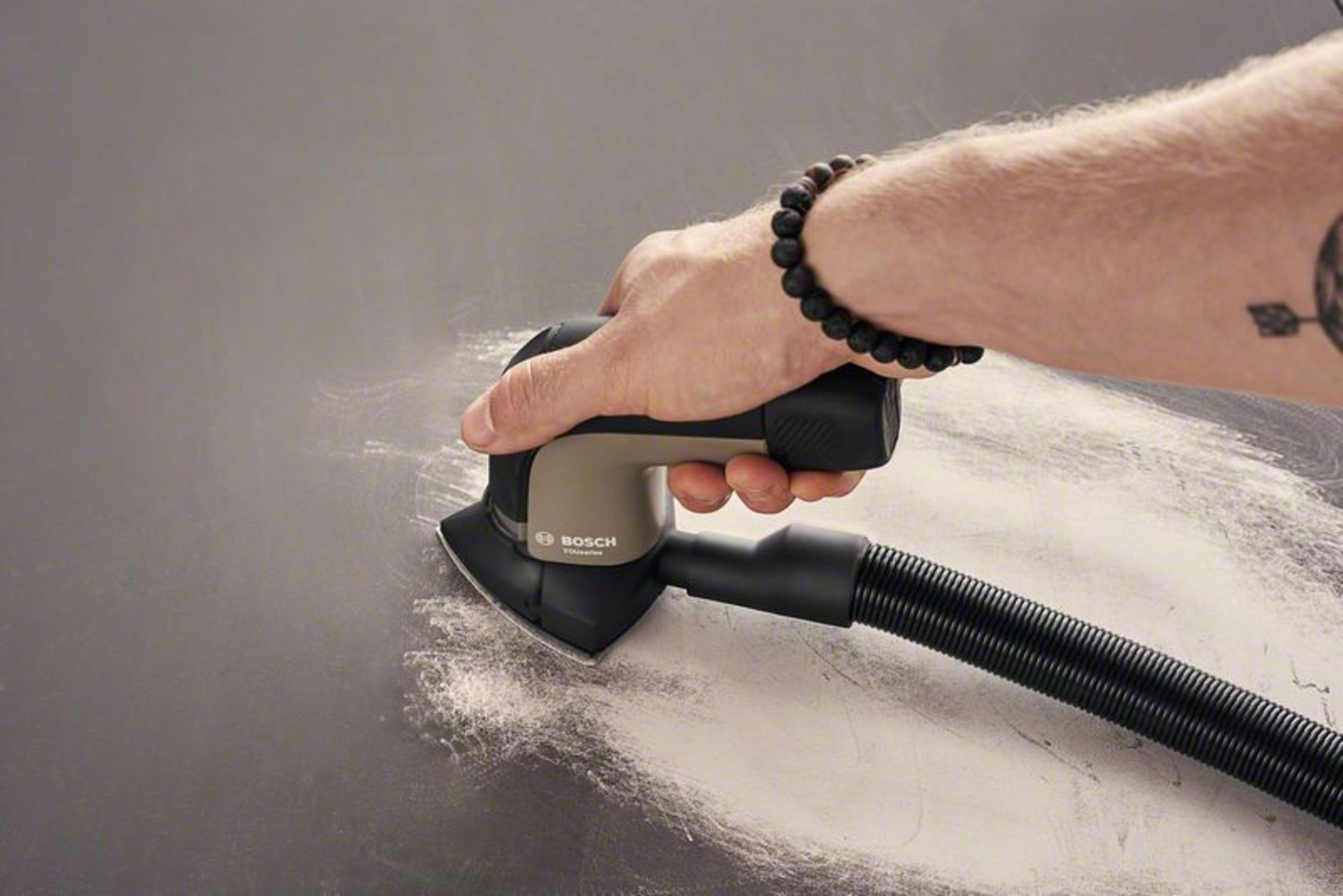 Eine Hand, die eine dunkle Oberfläche mit einem Bosch-Schleifer abschleift. Staub vorhanden.