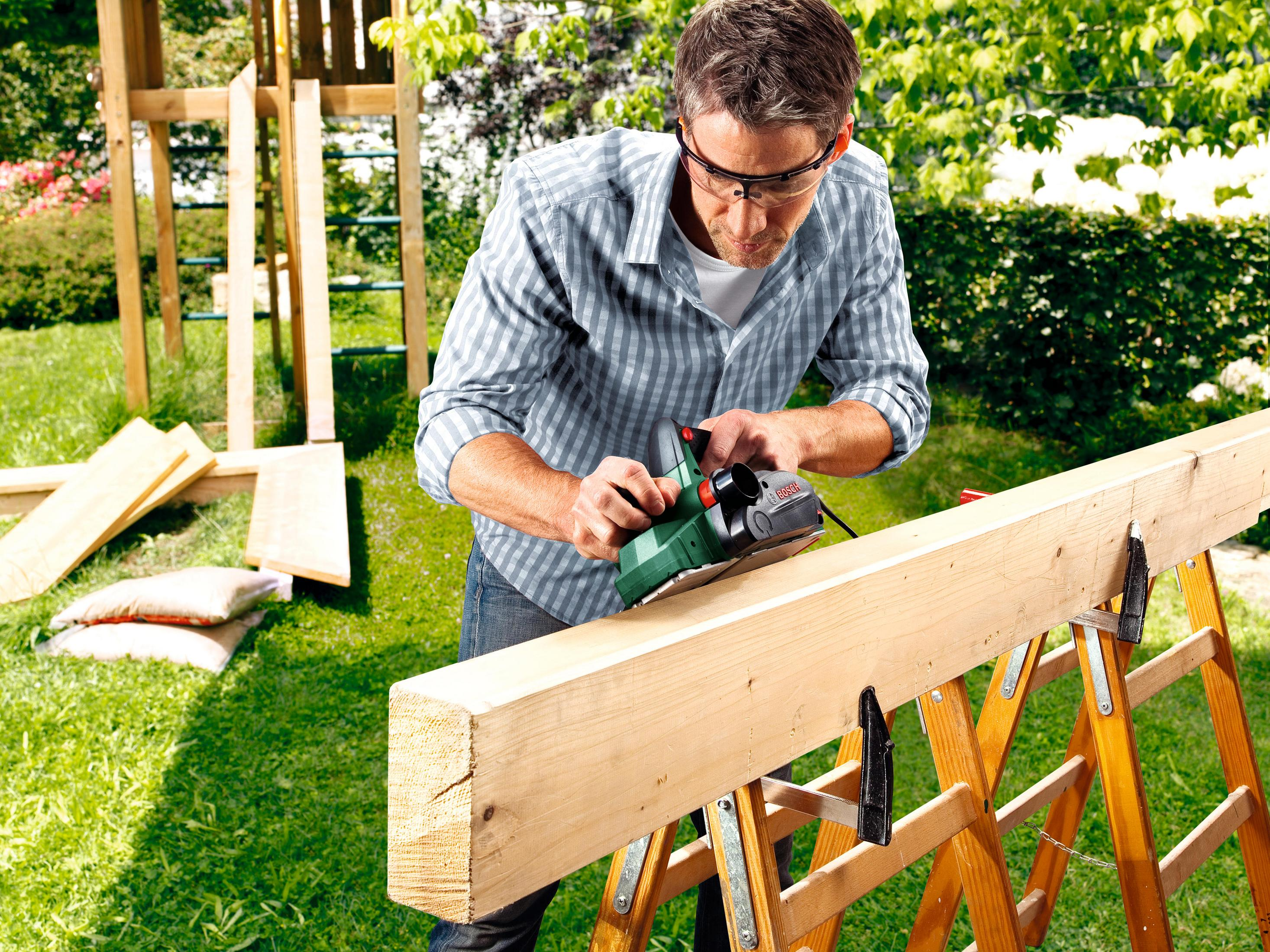 Un uomo che usa una pialla Bosch su una trave di legno all'aperto, con un parco giochi sullo sfondo.