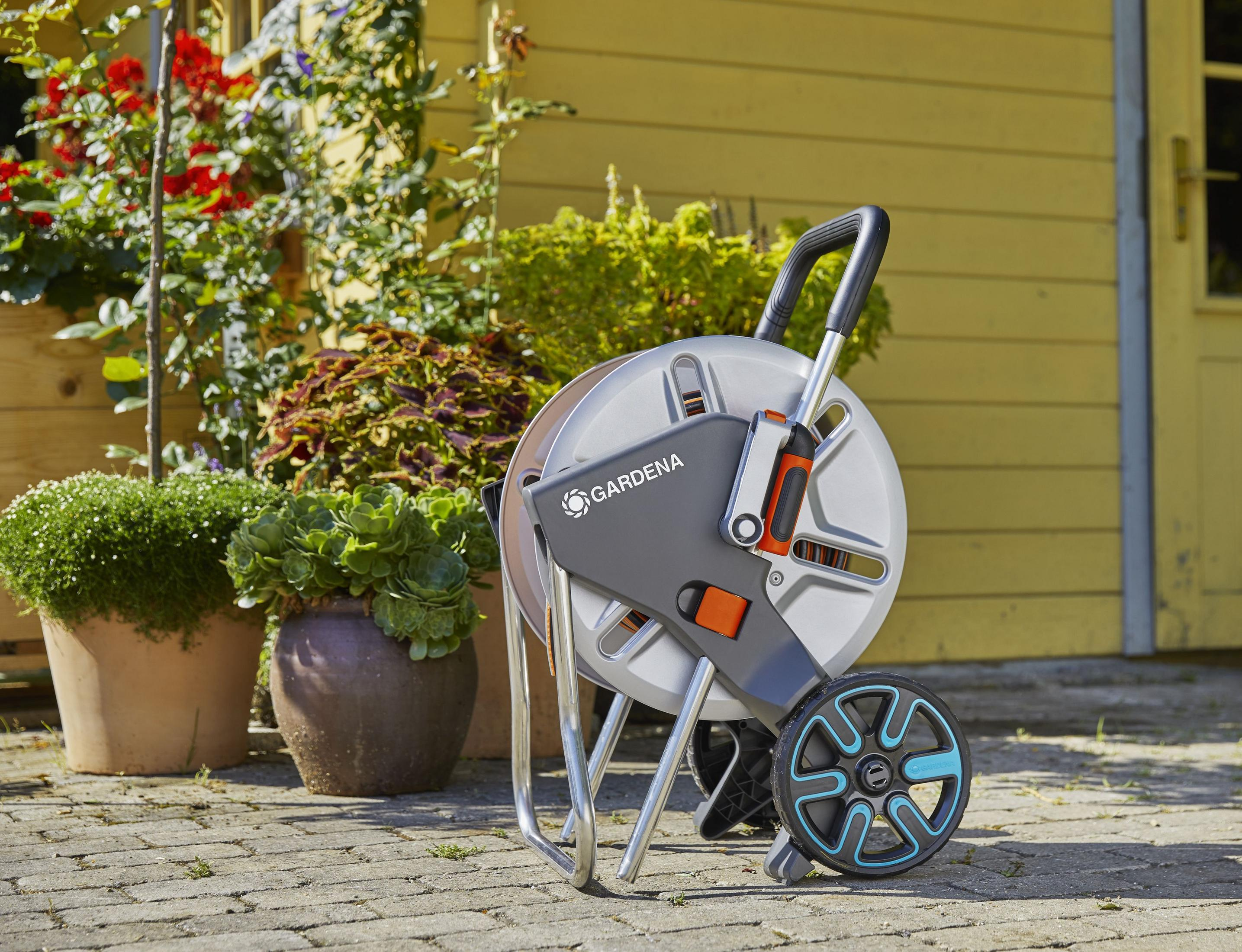 Un avvolgitubo da giardino Gardena grigio e nero con un tubo. Piante e una casa gialla sullo sfondo.