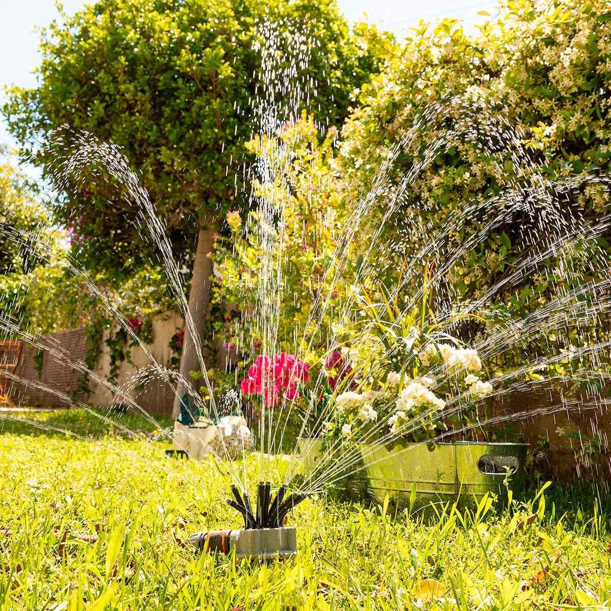 Sprinkler bewässert einen grünen Rasen. Bäume und Blumen sind im Hintergrund.