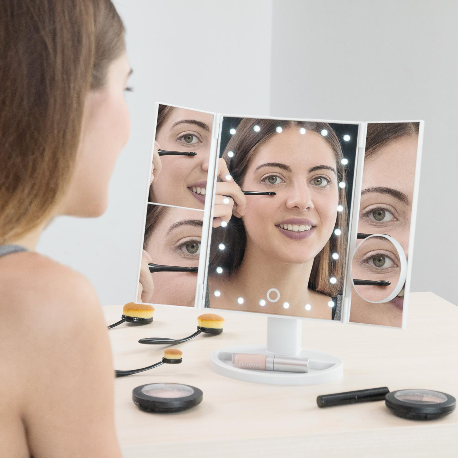 Una mujer usando un espejo de maquillaje con luces y brochas de maquillaje sobre la mesa.
