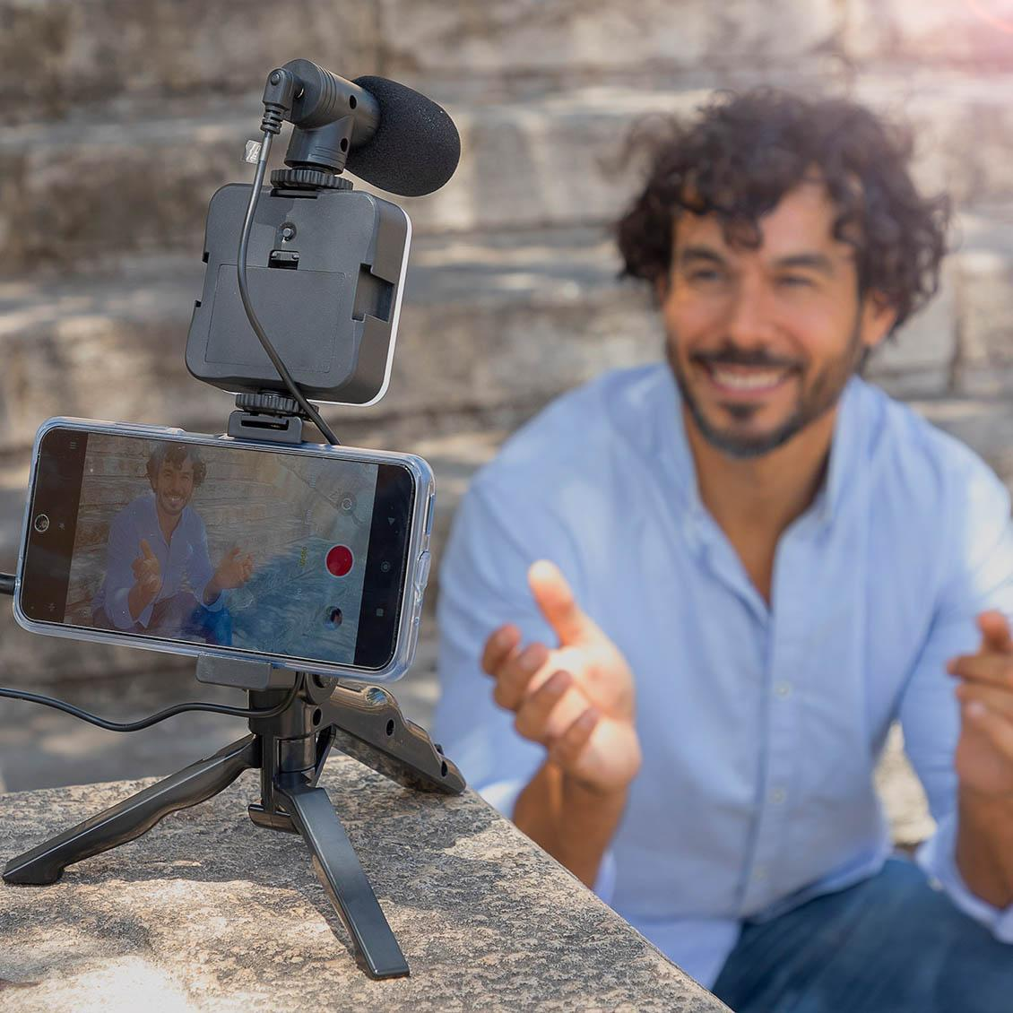 Un hombre se filma con un smartphone y un trípode, sonriendo a la cámara.