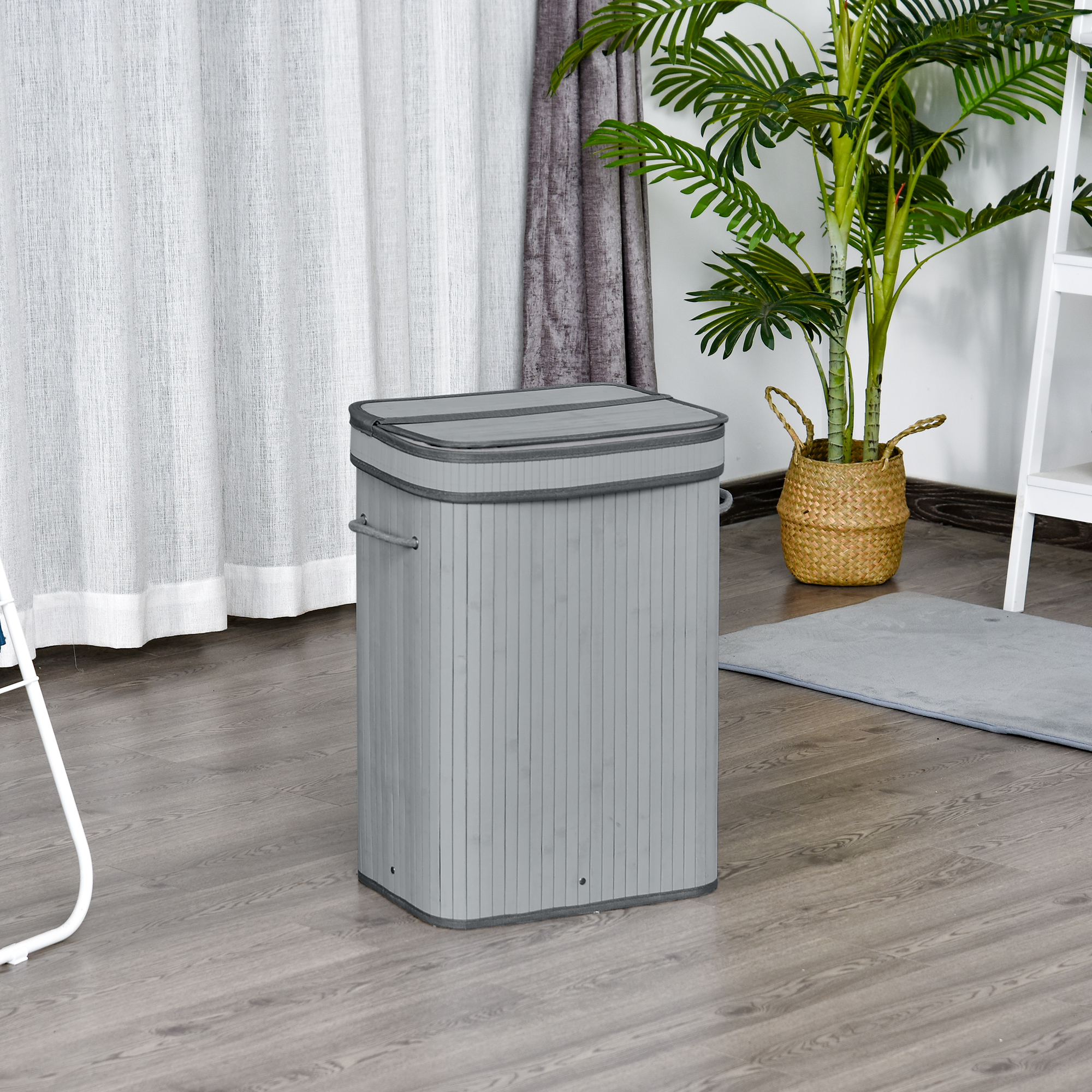 Grauer Wäschekorb. Ein weißer Vorhang, eine Pflanze und ein kleiner Teppich im Hintergrund.