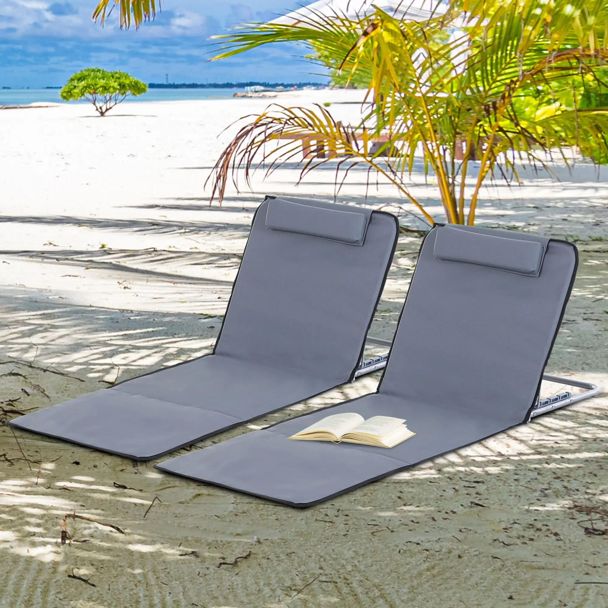 Zwei graue Strandstühle an einem Sandstrand mit einem Buch. Palmen im Hintergrund unter sonnigem Himmel.