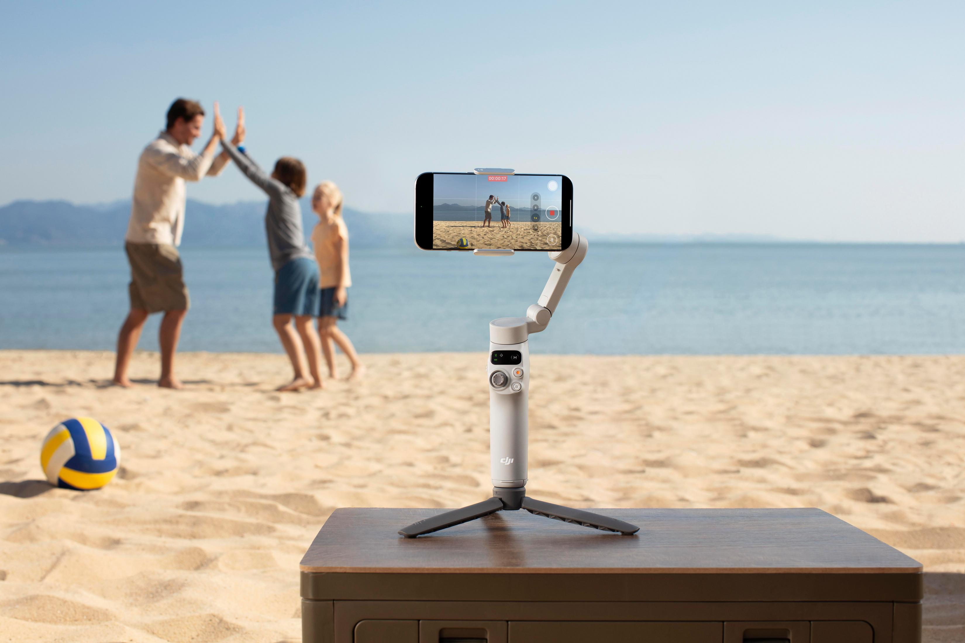 Telefon na statywie nagrywa rodzinę na plaży. W tle piłka do siatkówki i ocean.