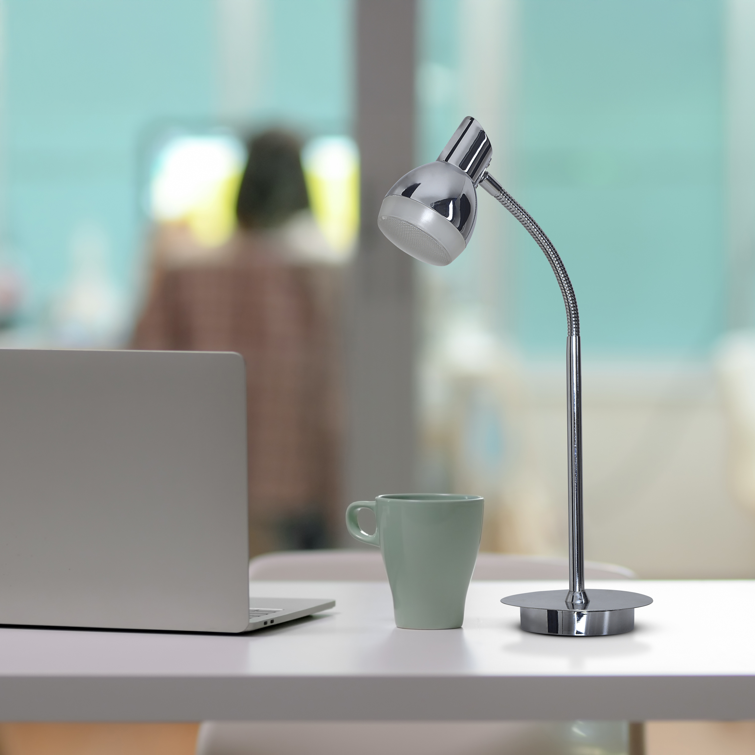 Eine silberne Schreibtischlampe auf einem weißen Schreibtisch. Ein Laptop, eine grüne Tasse und unscharfer Hintergrund.