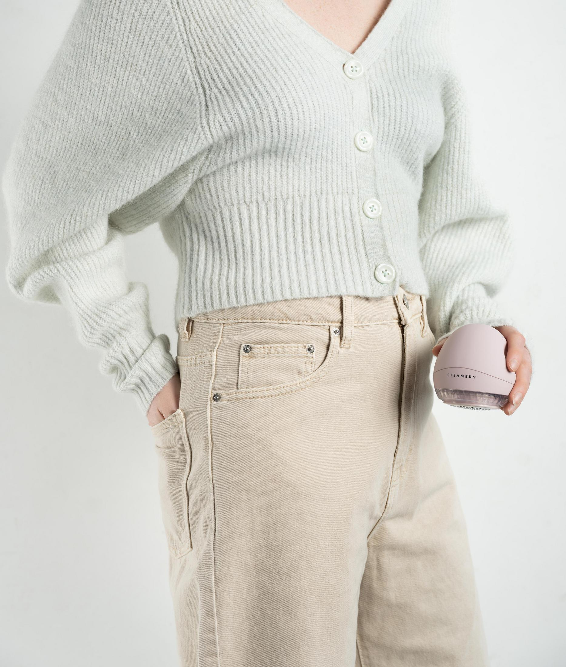 Eine Person hält ein rosa Dampfgerät, trägt einen Pullover und Jeans. Weißer Hintergrund.