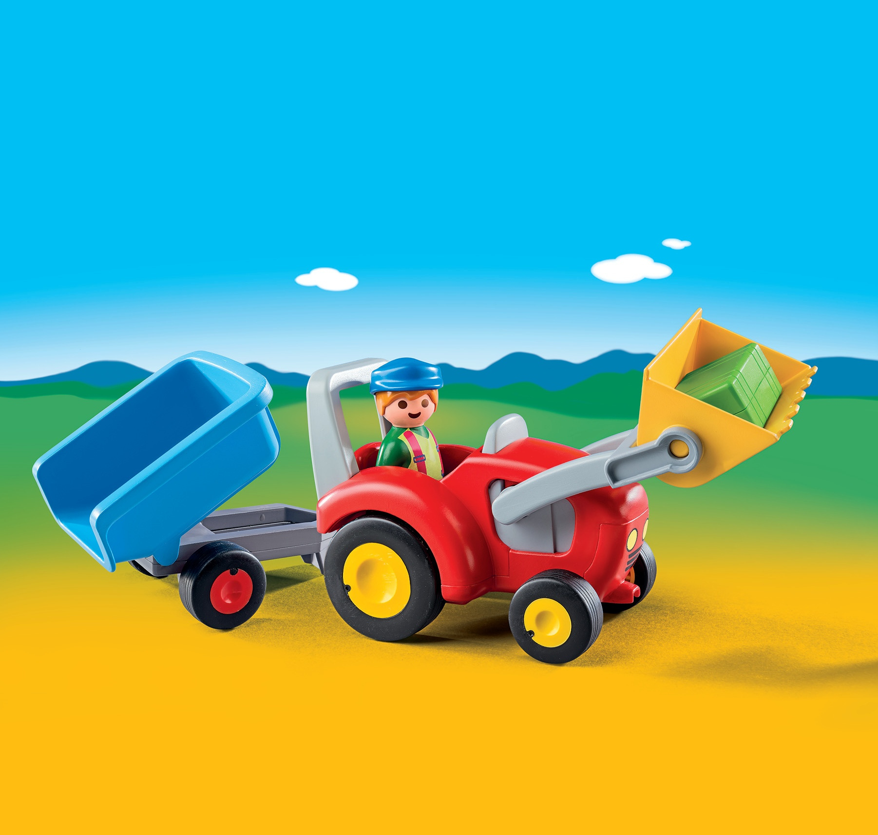 Ein roter Spielzeugtraktor mit blauem Anhänger und gelber Schaufel, gefahren von einer Spielfigur unter blauem Himmel.