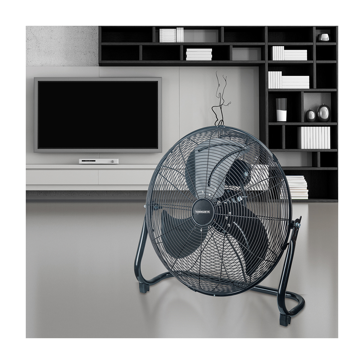 Un ventilador negro grande en una habitación, frente a un televisor, con una estantería negra en la pared.