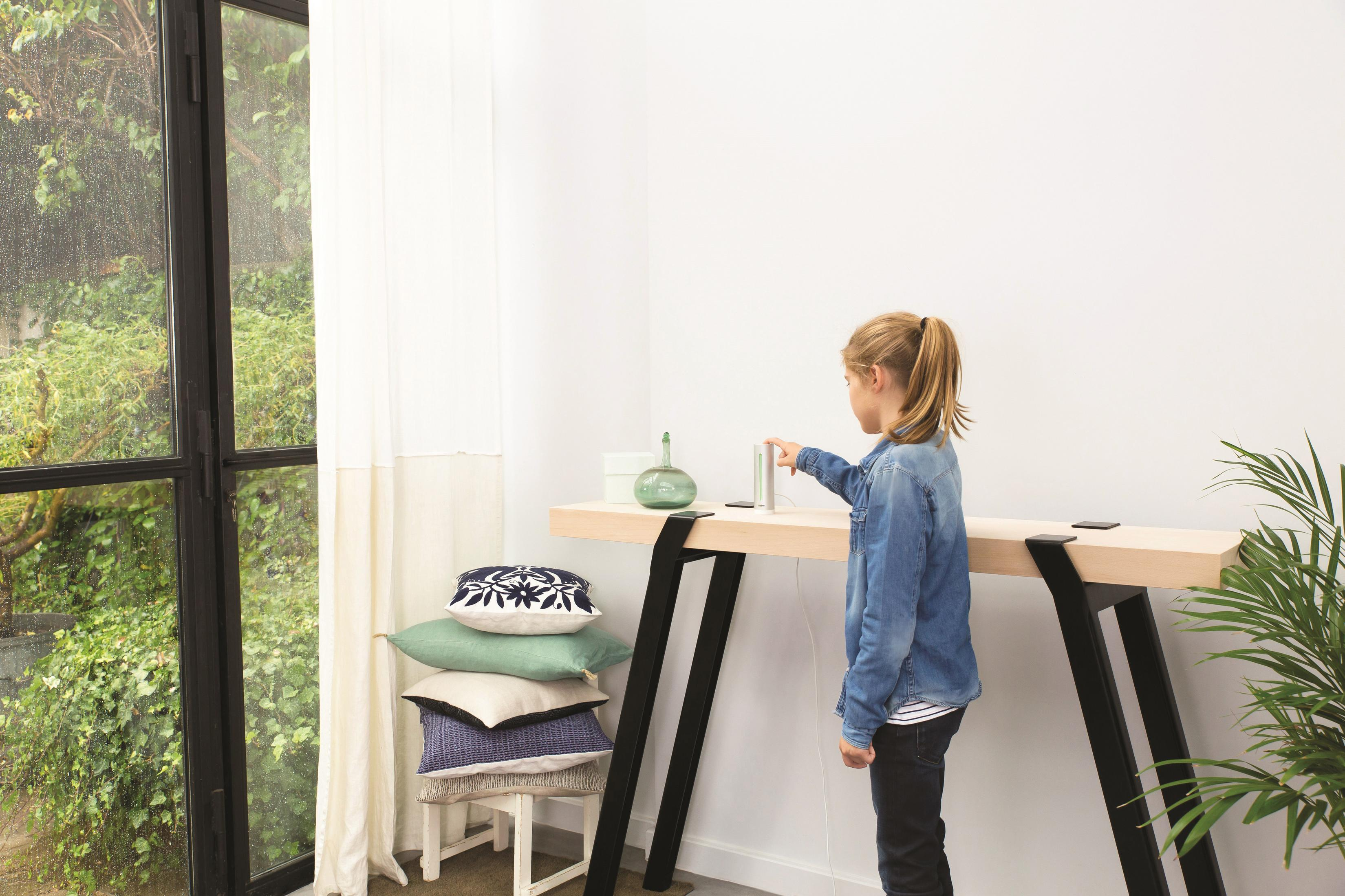 Dziewczyna dotyka urządzenia na stole. Stół przy oknie z zasłoną. Poduszki i roślina na pierwszym planie.
