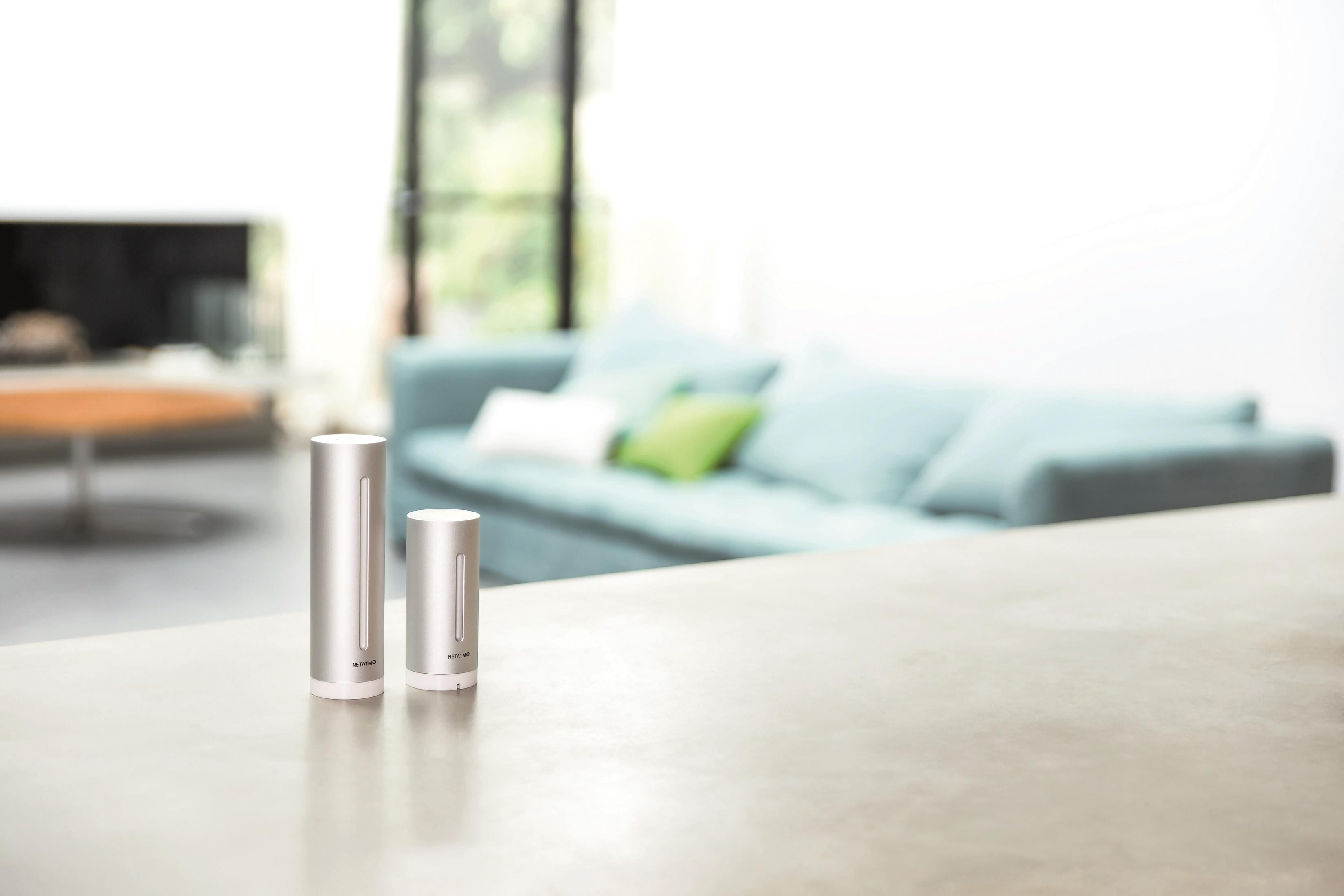 Dwa srebrne urządzenia smart home na błyszczącej powierzchni, z rozmytym tłem salonu.