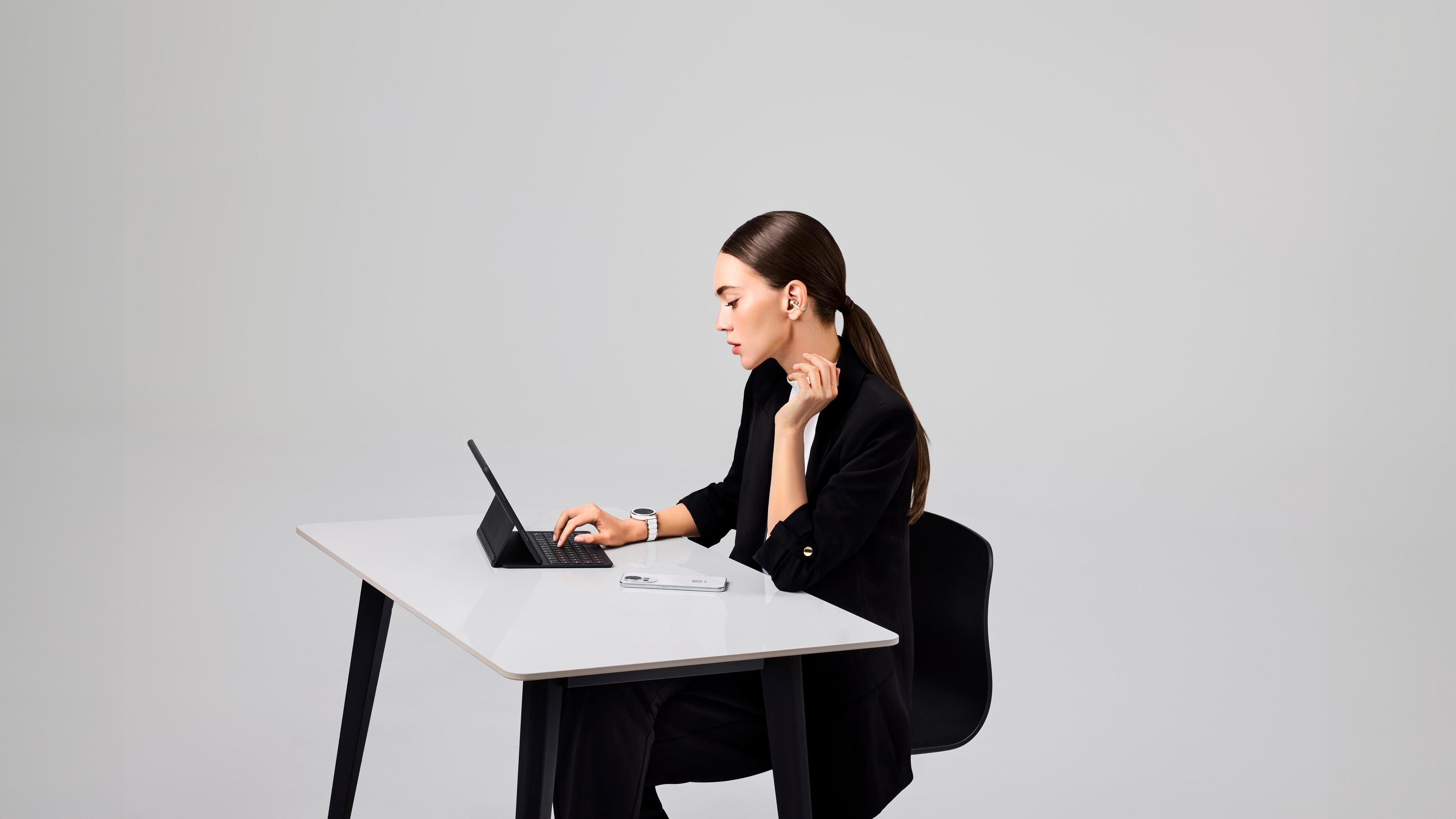 Kobieta w czarnym garniturze pracuje przy białym biurku, używając tabletu. Ma włożoną słuchawkę.