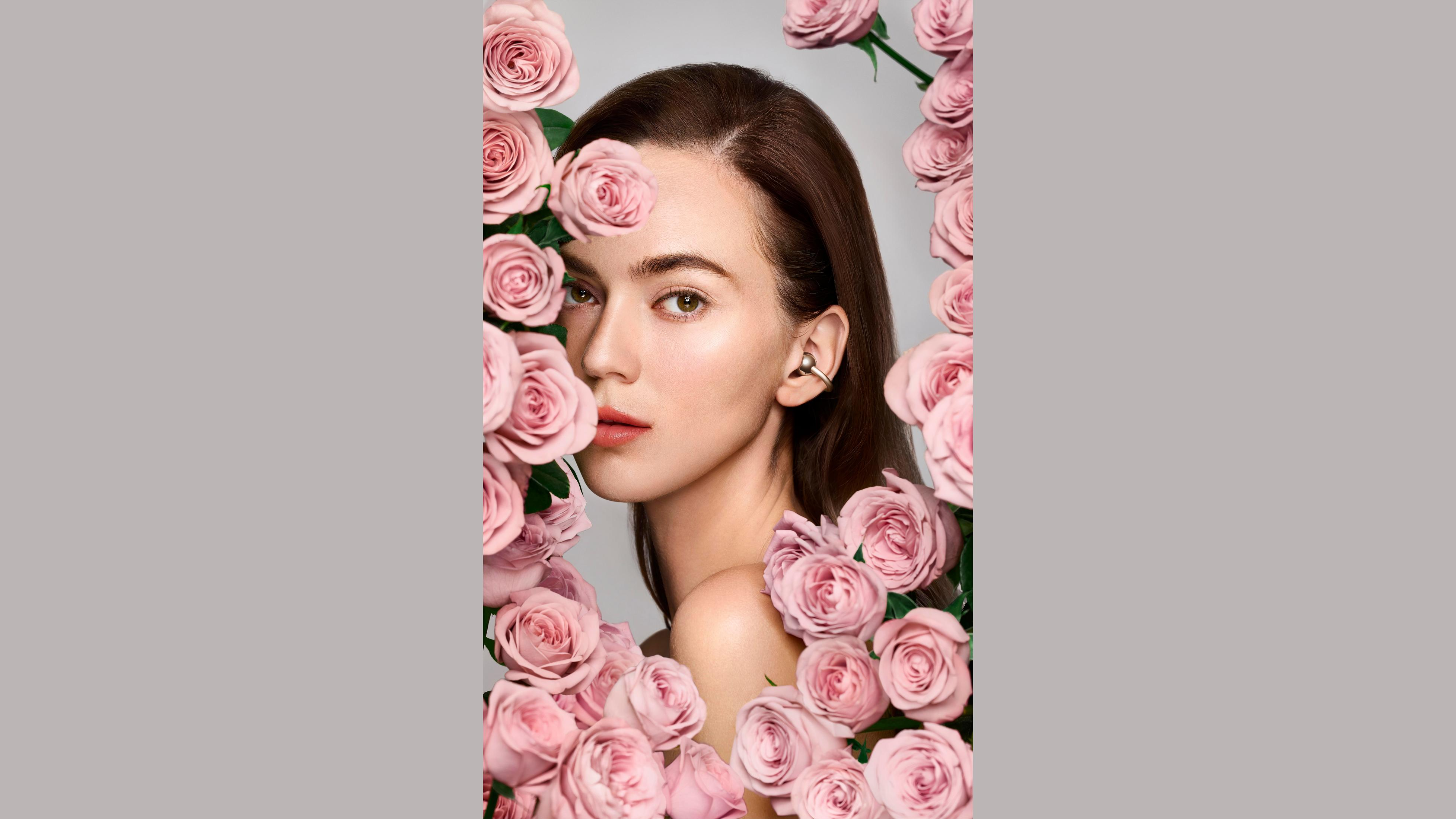 Twarz kobiety otoczona różowymi różami. Nosi kolczyki. Tło jest szare.