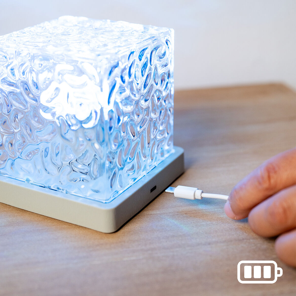 Ein leuchtender, strukturierter Glaswürfel auf grauem Sockel, mit Hand, die Ladekabel einsteckt.