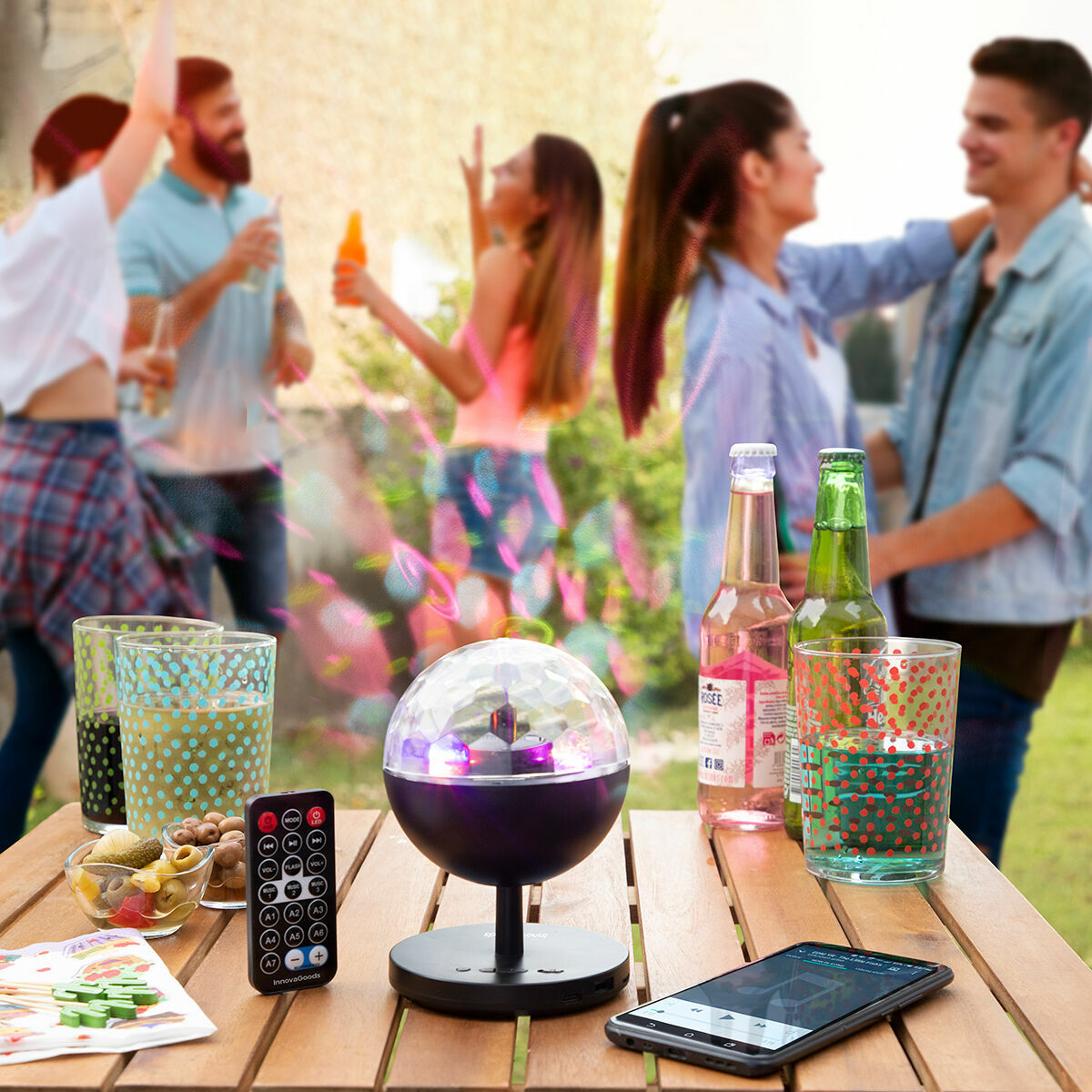 Partyszene: Lautsprecher, Lichter, Getränke, Telefon, tanzende Leute im Hintergrund. Holztisch, Outdoor-Umgebung.